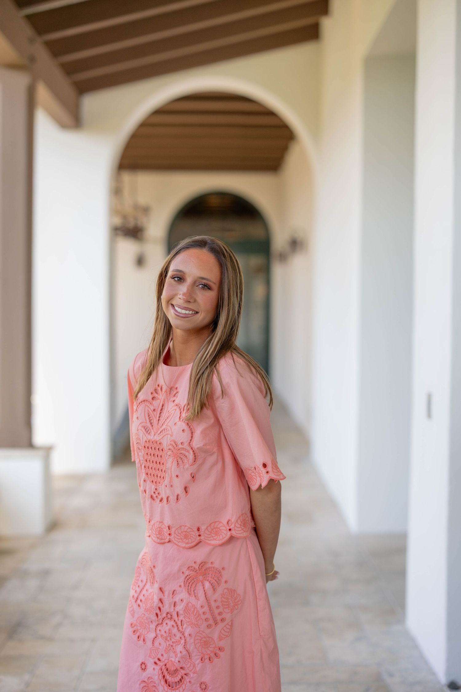 Person in coral pink dress poses along a covered walkway with arched columns.