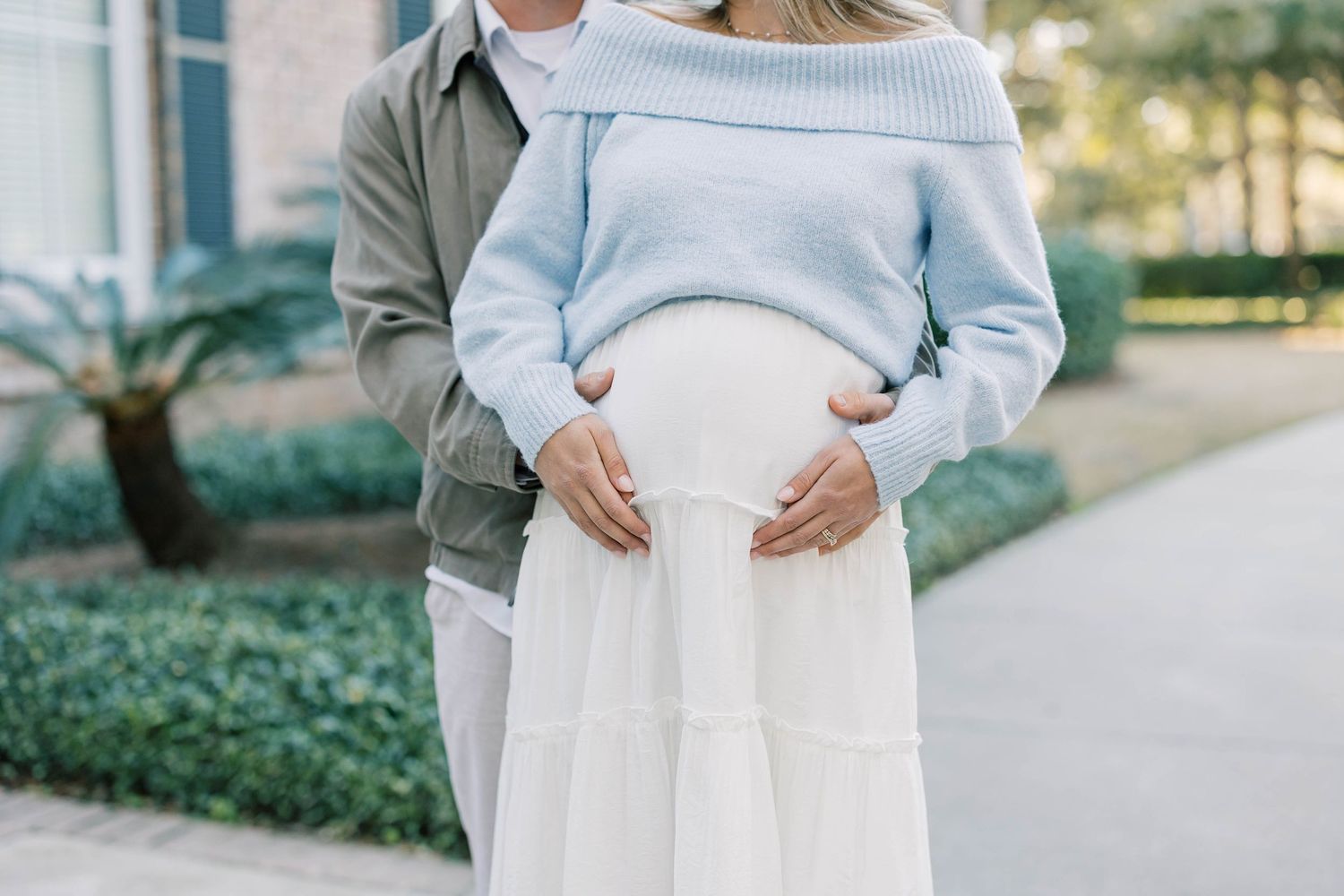 Couple cradling pregnant belly outdoors, woman in blue sweater and white skirt, man in olive jacket.