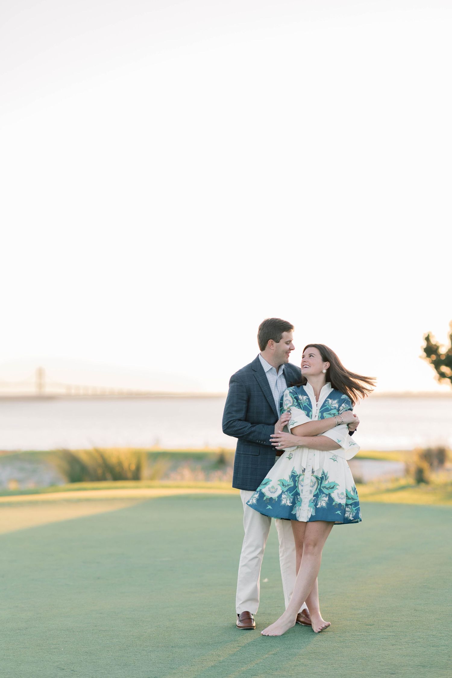 Couple embracing on green lawn with bridge backdrop at golden hour, woman in floral dress, man in navy blazer.
