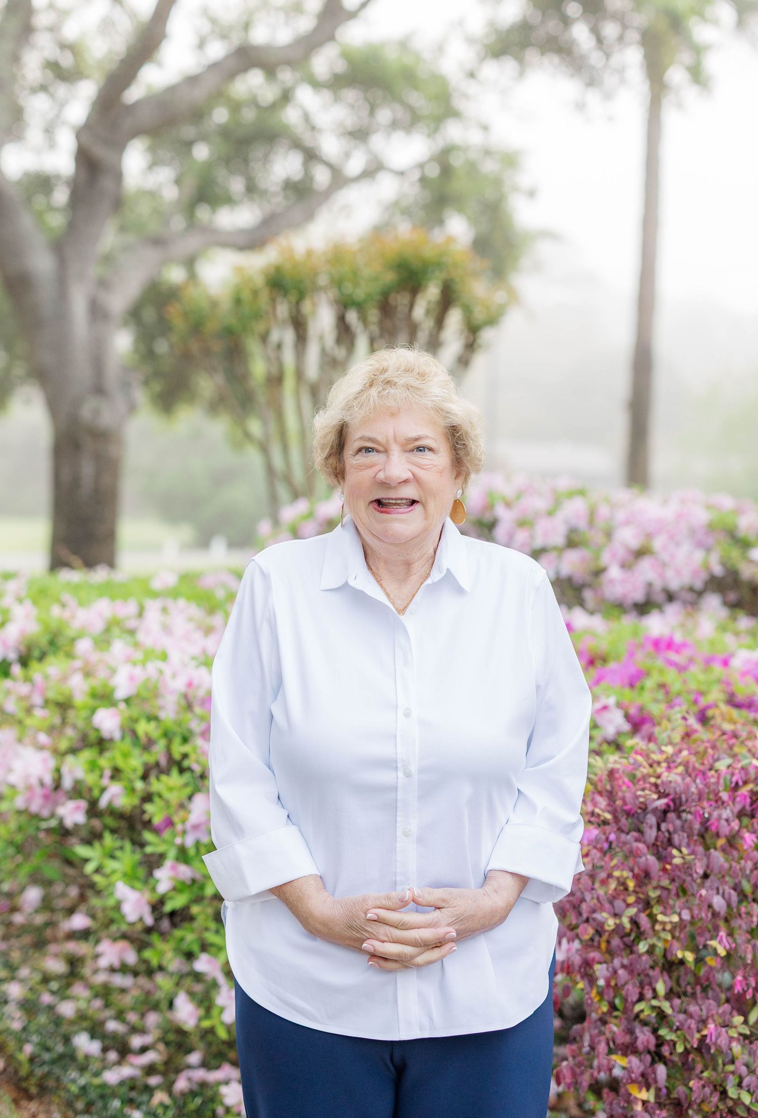 Smiling older woman in white shirt standing among colorful pink flowering bushes in a lush garden setting.