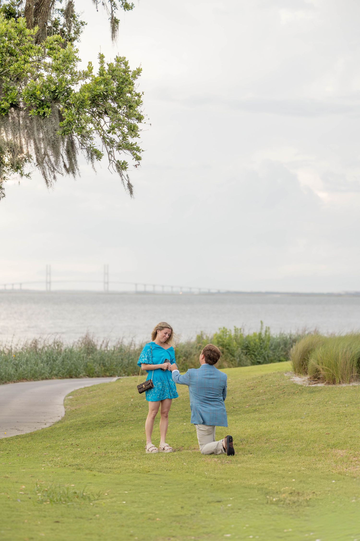 Man proposes on one knee to woman in teal dress by waterfront park with bridge in background.