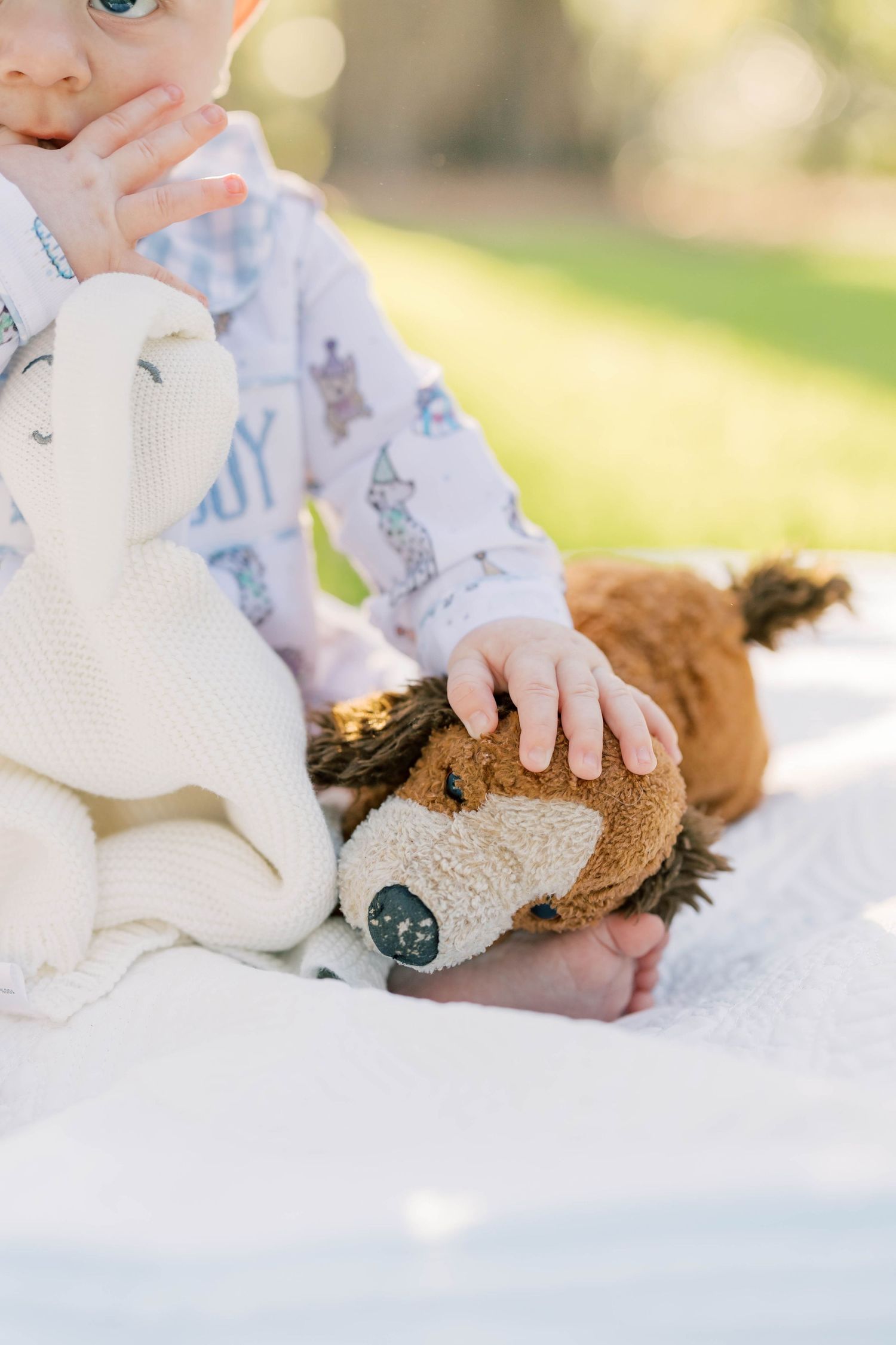 Baby in bear pajamas sits on white blanket outdoors, holding a plush dog toy with a cream knit blanket beside them.