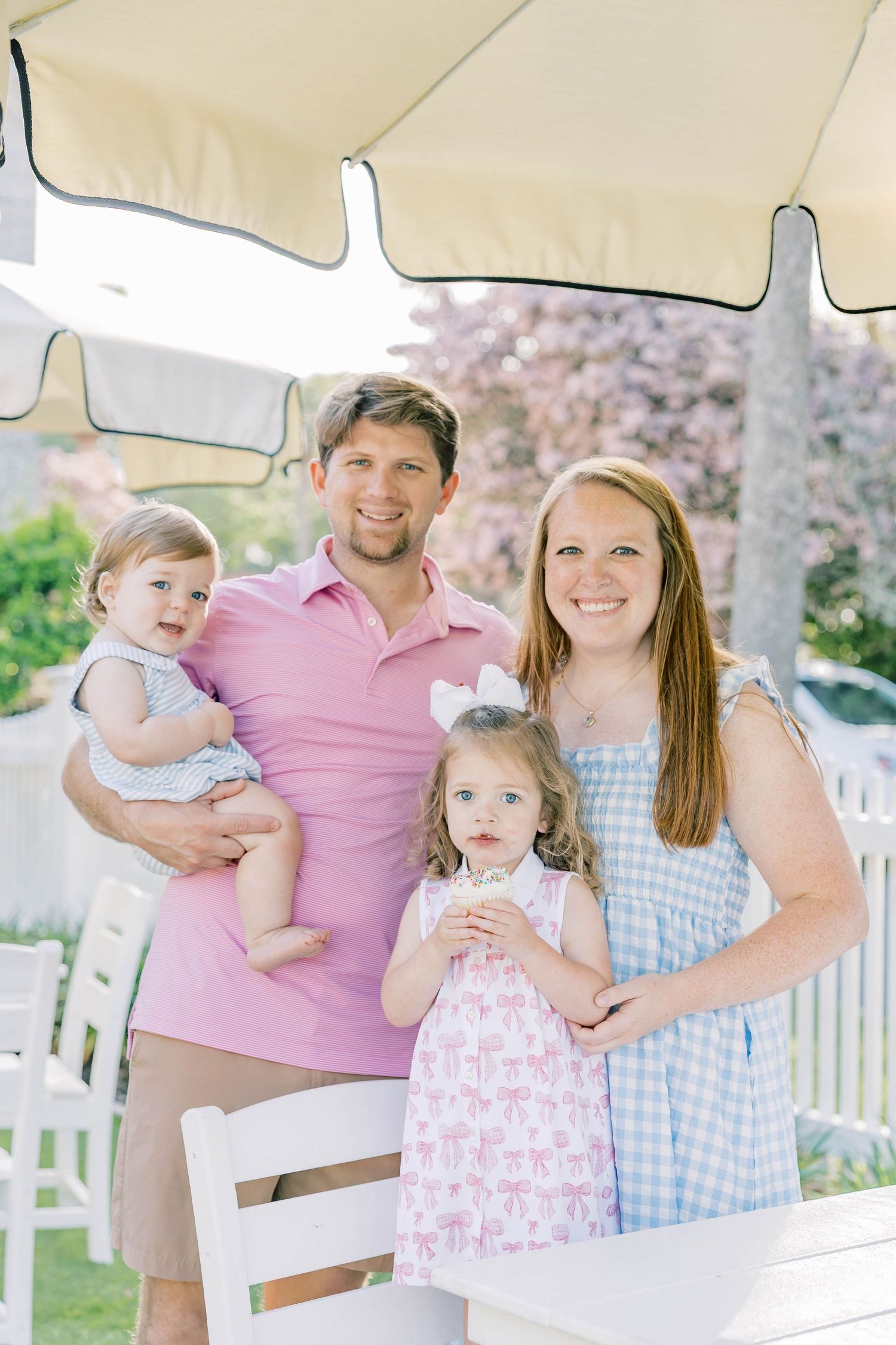 A smiling family of four poses outdoors under a striped umbrella, with parents holding two young daughters in pastel outfits.