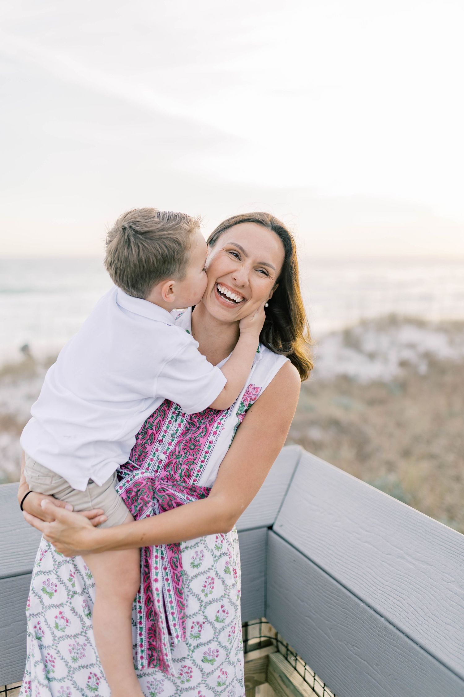 A joyful mother laughs as her young son kisses her cheek on a beachside deck at sunset.