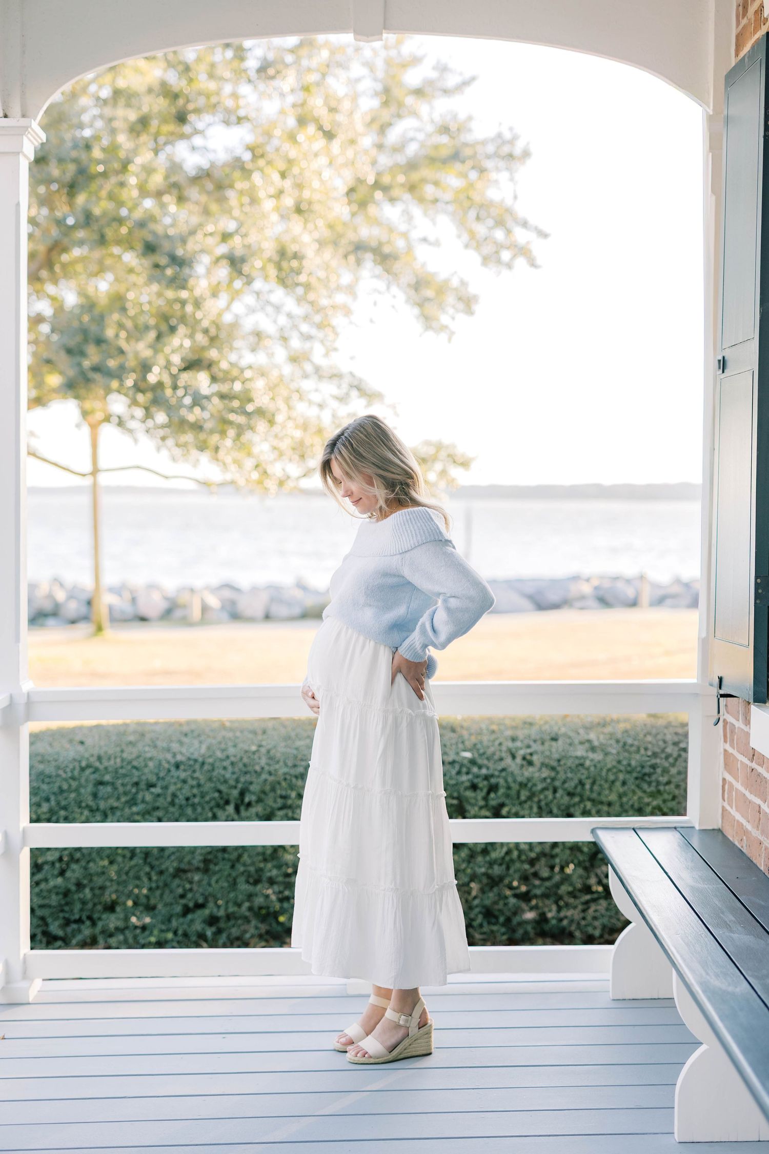 Pregnant woman in white dress and blue top poses on a waterfront porch in soft natural light.