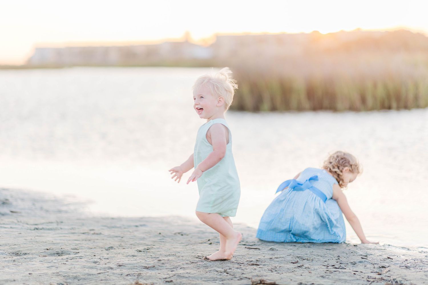 Two toddler girls playing on a sandy beach at golden hour, one laughing and running in a mint romper.