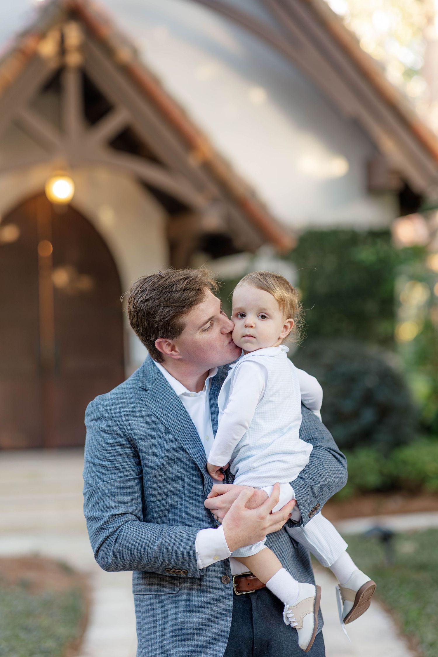 A man in a gray blazer kisses a toddler in white outside a charming chapel.