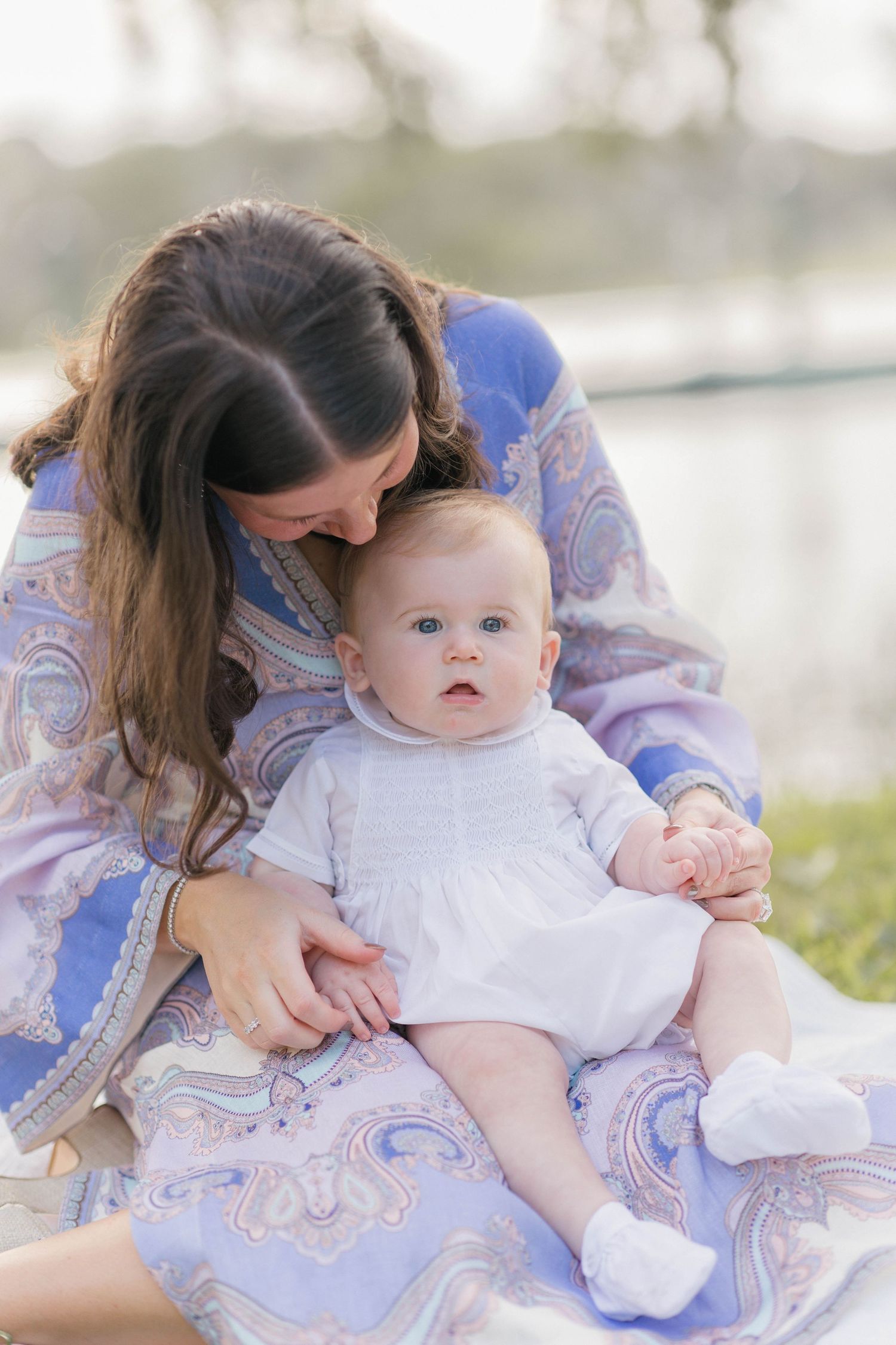 Mother in blue paisley dress kissing baby girl in white dress sitting outdoors near water.