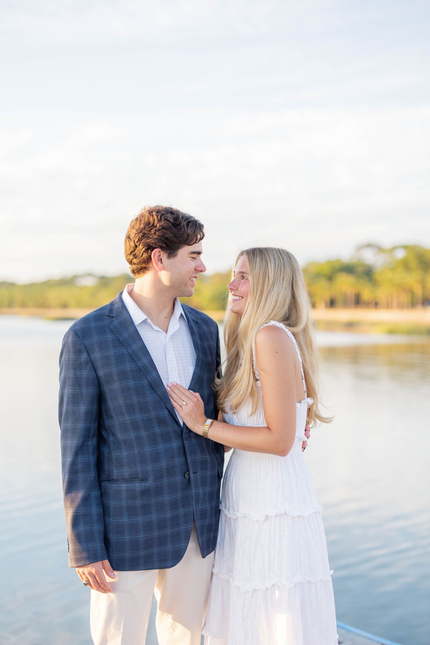 Happy couple posing near waterfront, man in plaid blazer and woman in white dress, smiling at each other.