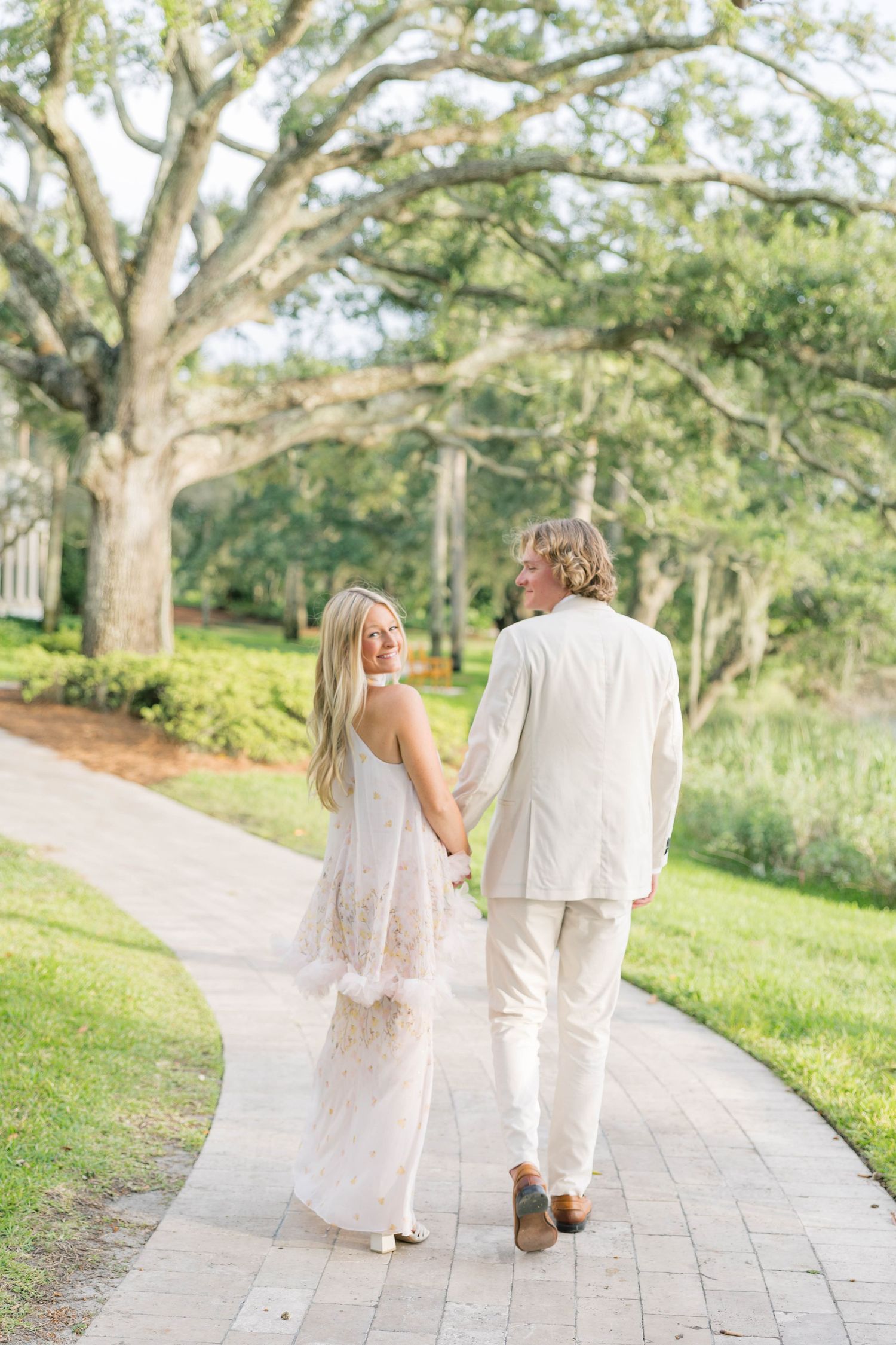 A couple in white wedding attire walks hand-in-hand along a brick path surrounded by lush green trees.