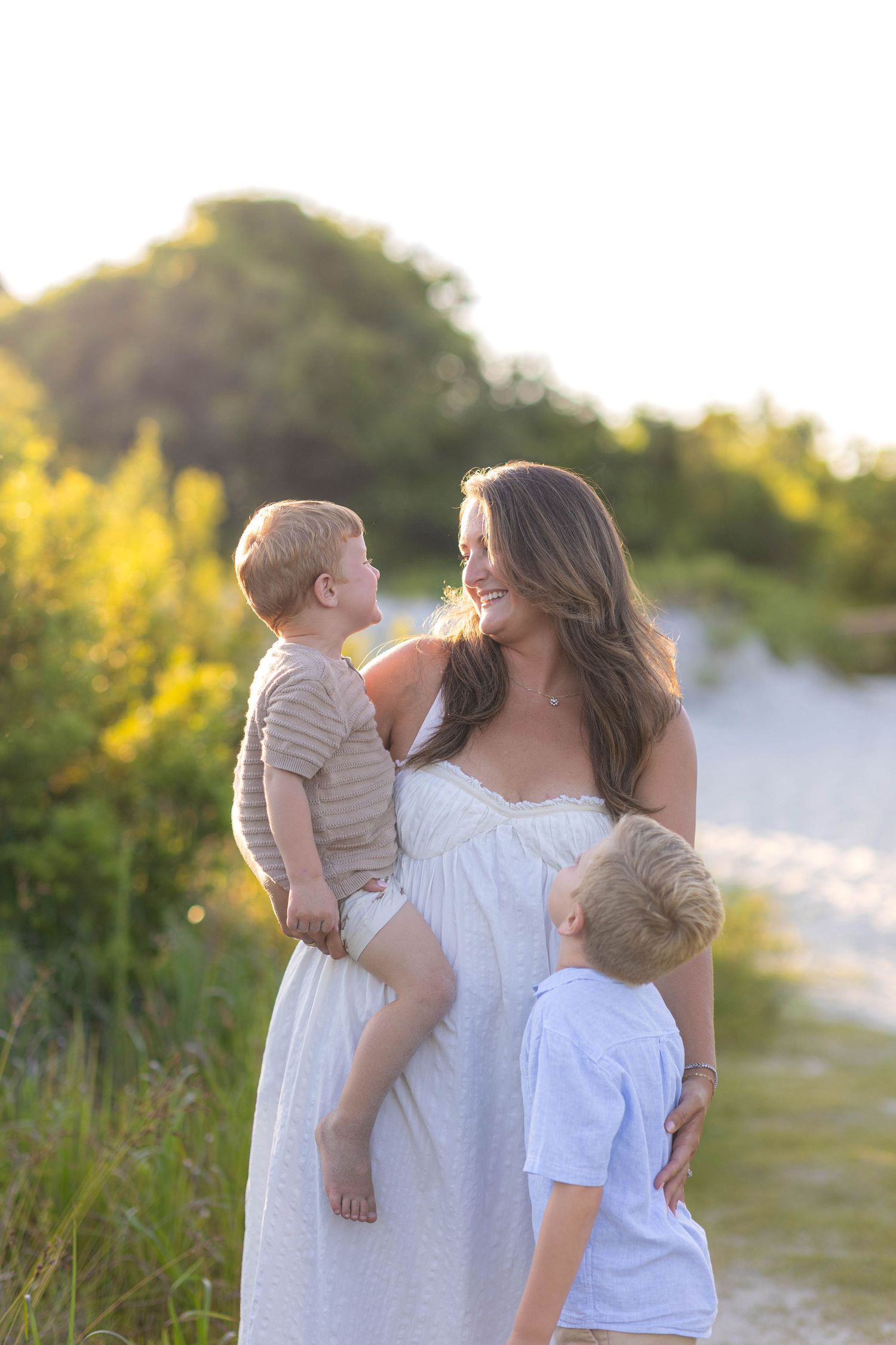 A pregnant mother holds two young boys outdoors in golden sunset light.