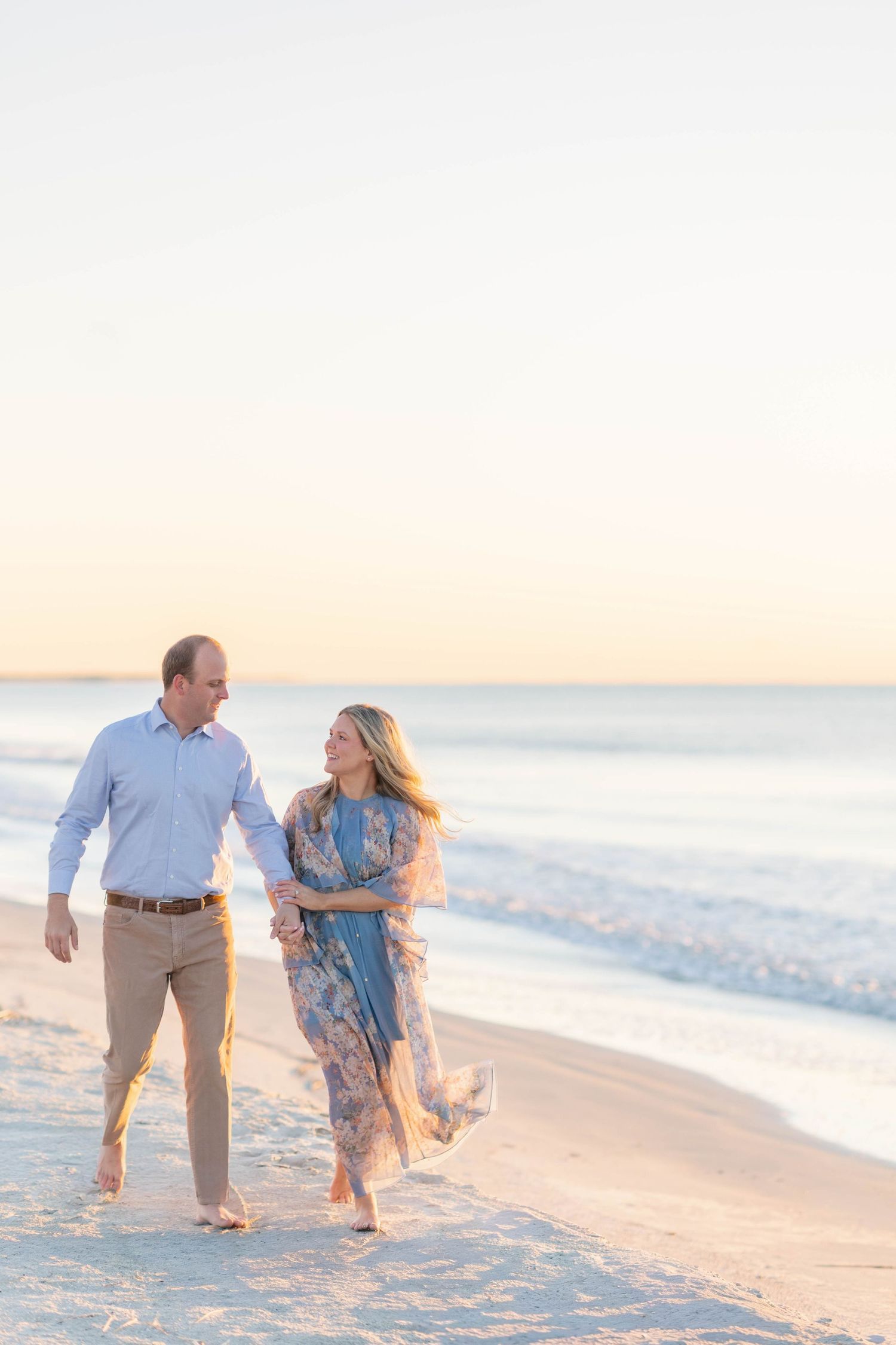 Couple walking hand in hand on a beach at sunset, woman in floral dress, man in light blue shirt and khaki pants.