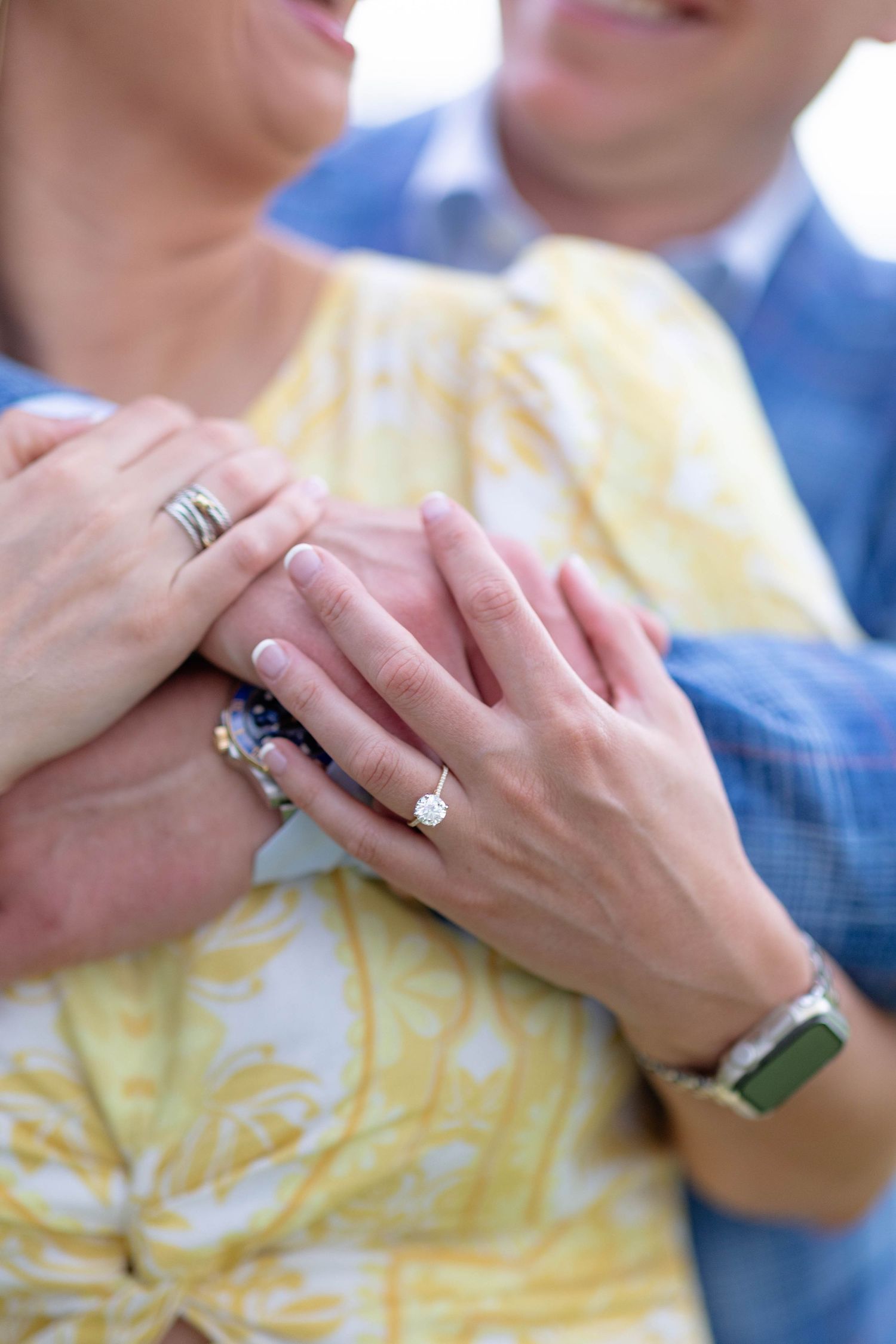 Close-up of couple's hands showing a diamond engagement ring, wedding band, and green smartwatch on a yellow dress.