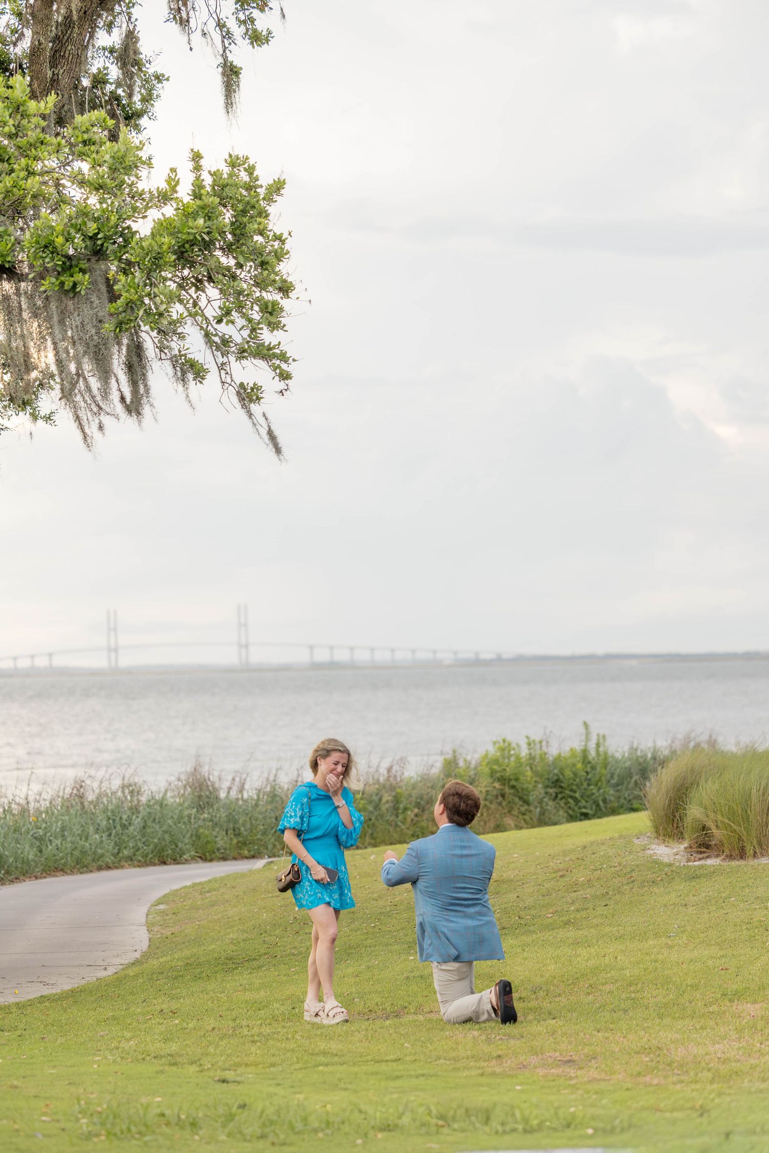 Man proposes on one knee to woman in blue dress by scenic waterfront with bridge views on cloudy day.
