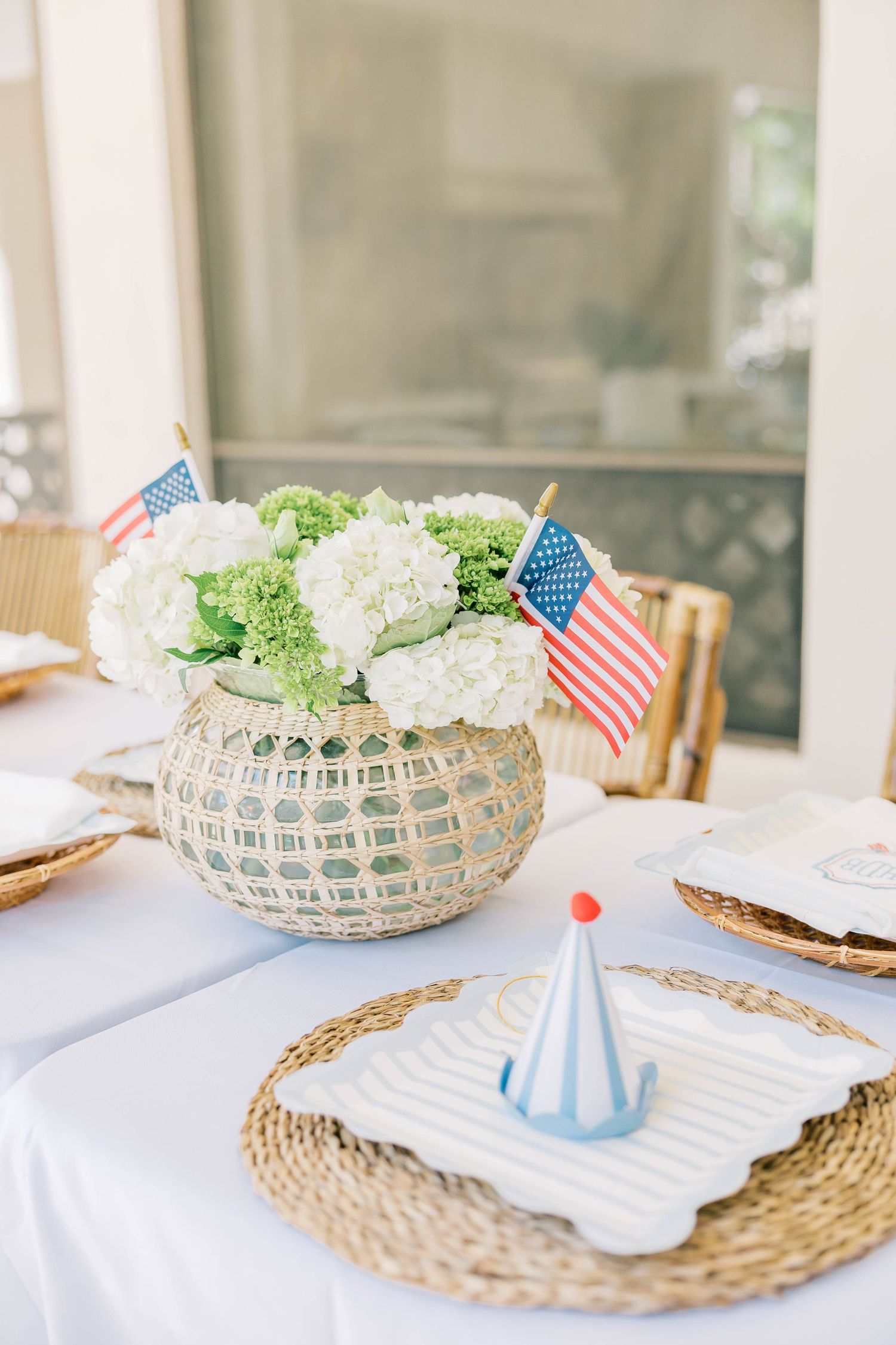 Patriotic 4th of July table setting with white hydrangeas, American flags, wicker chargers, and a party hat centerpiece.