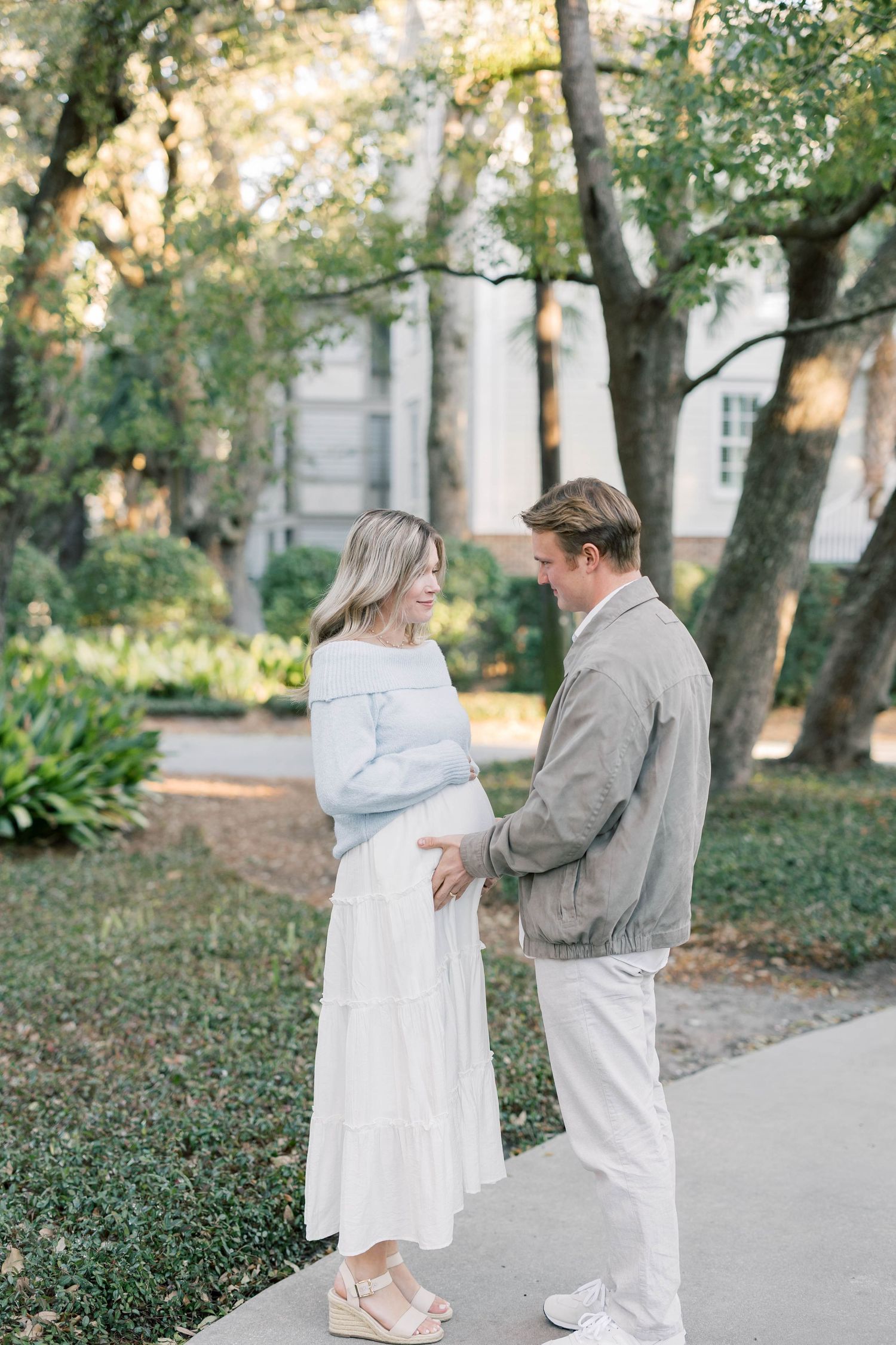 A couple shares a tender moment on a garden path, with the woman in a blue top and white skirt cradling her baby bump.