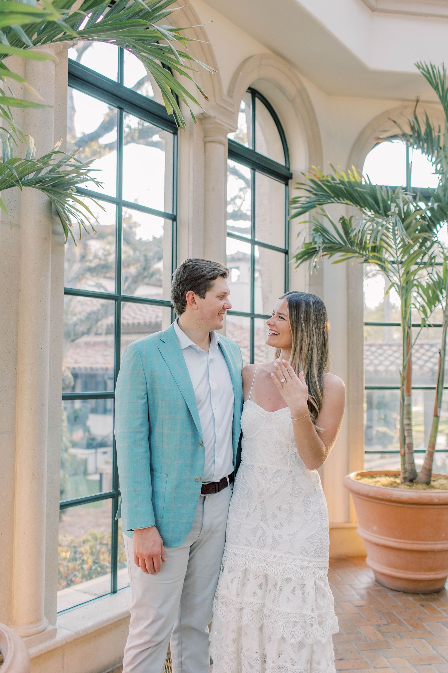 Couple smiling at each other in elegant sunroom with arched windows, man in light blue blazer, woman in white lace dress.