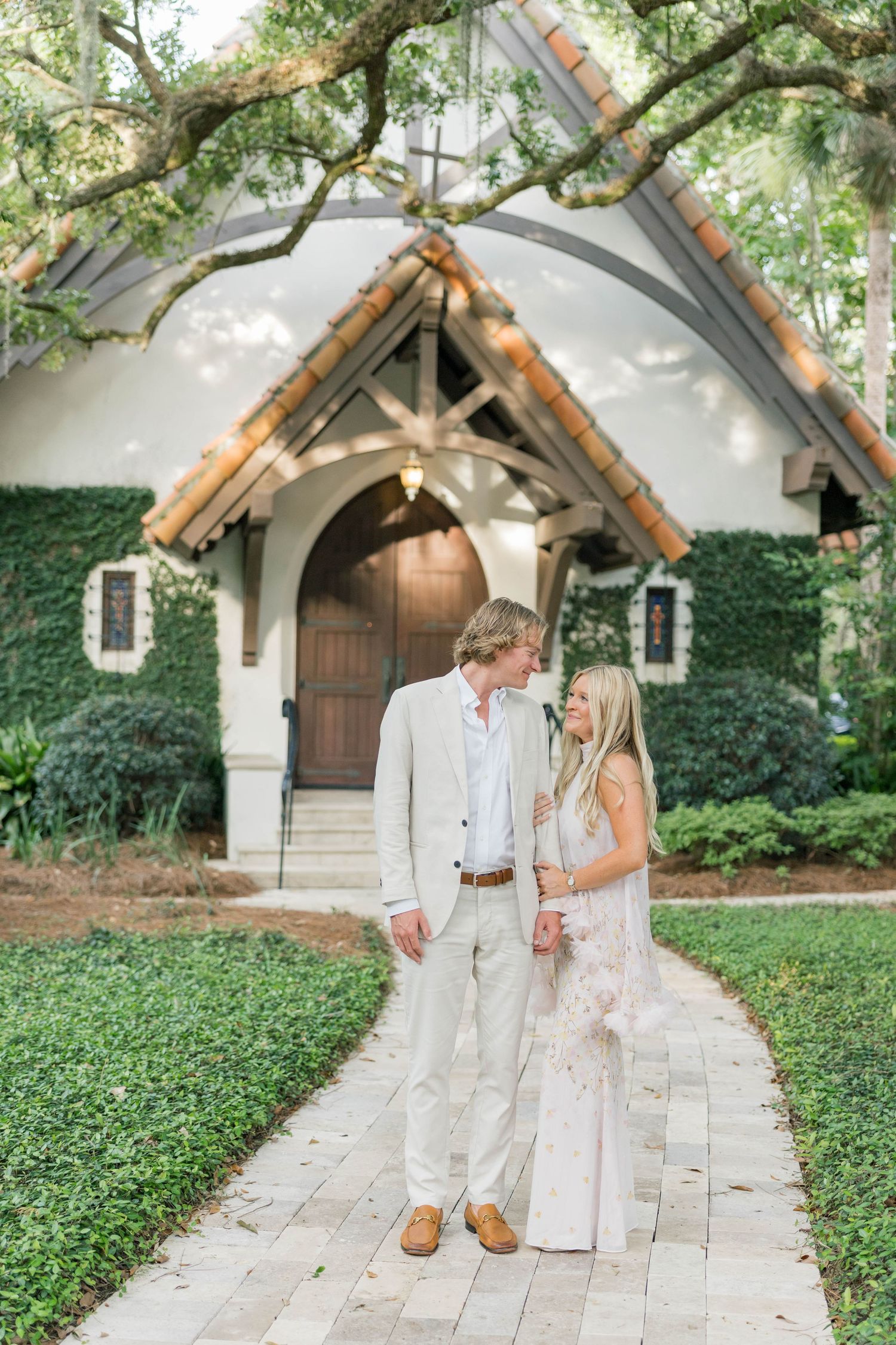 Couple in white attire holding hands on a brick path before a charming ivy-covered chapel surrounded by lush greenery.