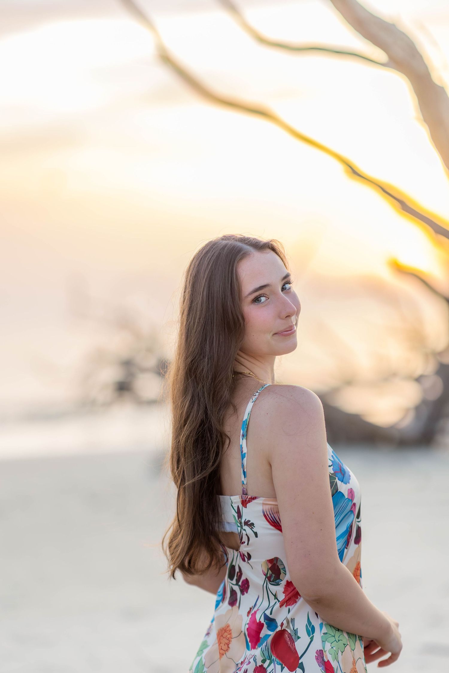 Young woman in floral sundress standing on beach at golden sunset with driftwood tree behind her.