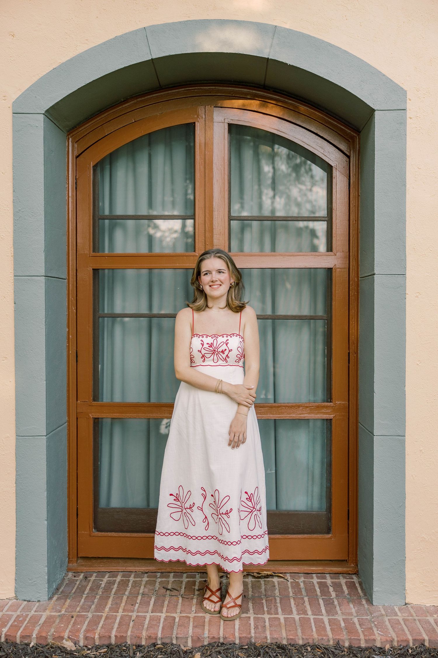 Young woman in white floral embroidered sundress standing before arched wooden door with blue trim.