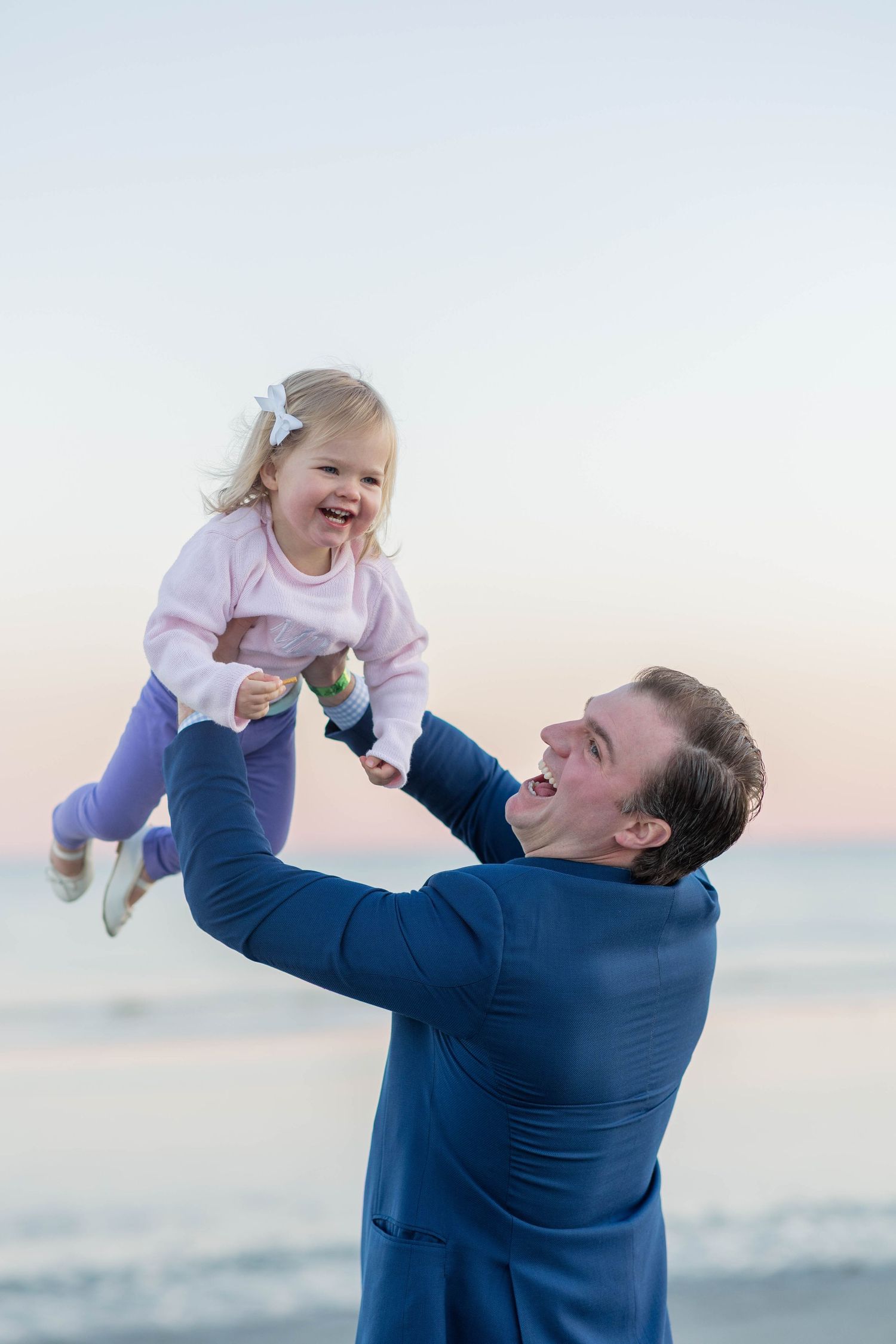Father joyfully lifting laughing toddler girl in the air on a beach at sunset.