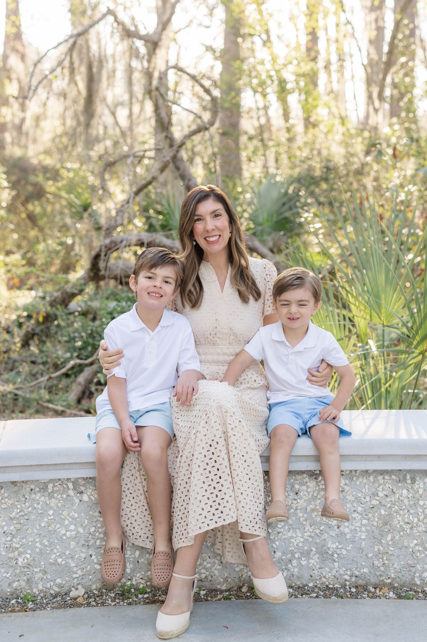 A smiling woman in a cream dress sits on a bench outdoors with two young boys in white shirts and blue shorts.