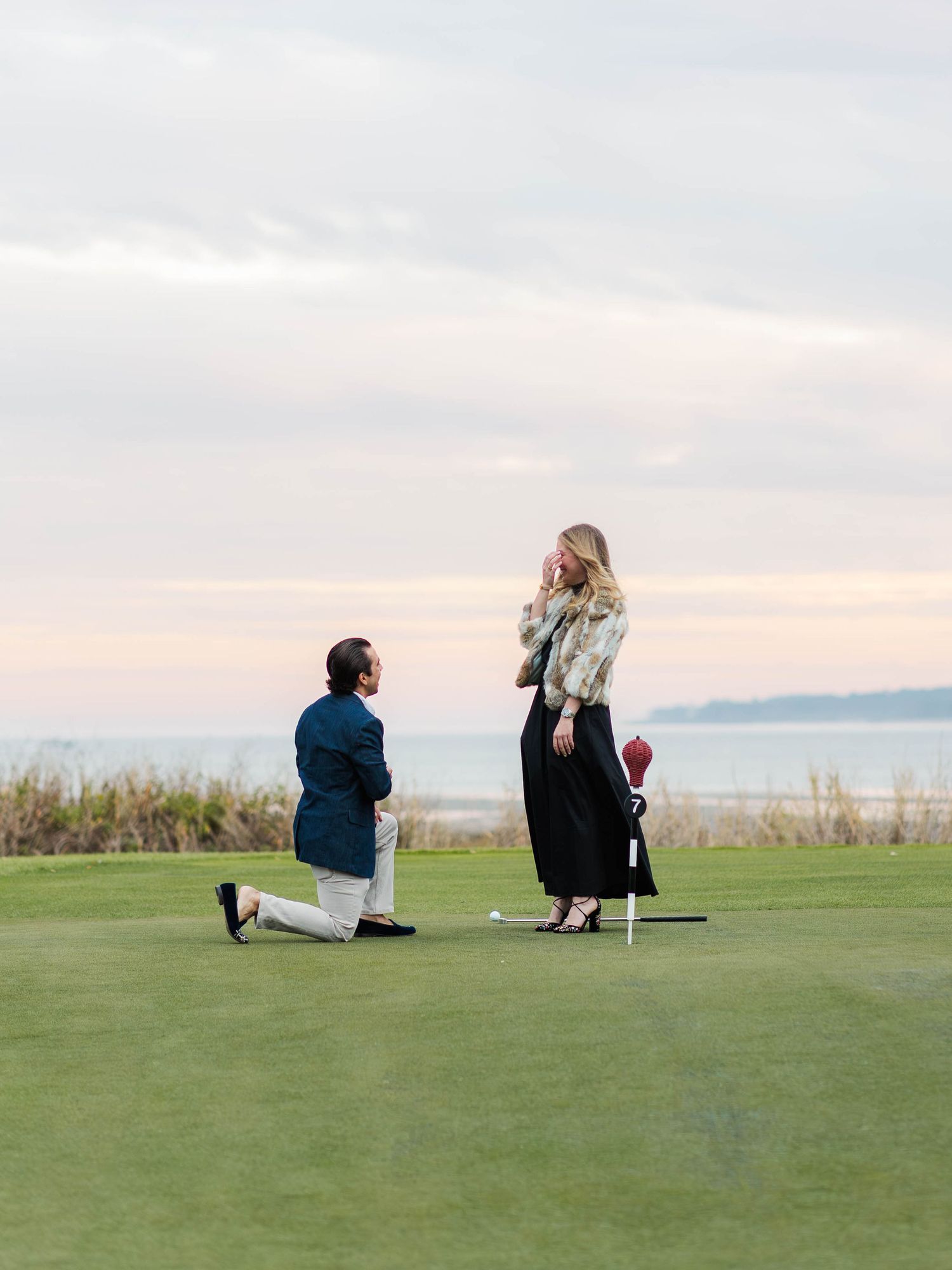 Man kneeling on golf course proposing to woman at sunset near ocean, romantic outdoor marriage proposal.
