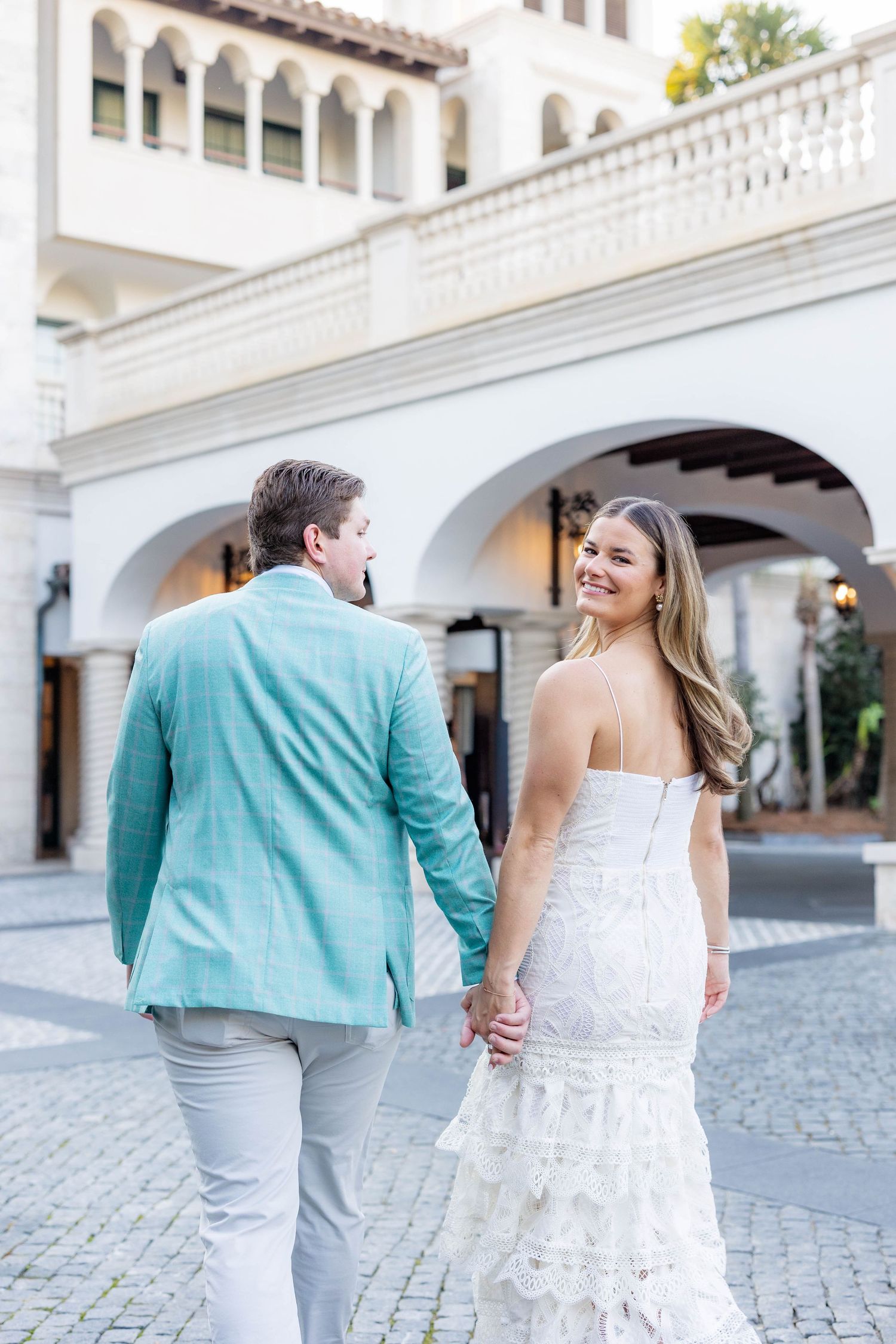 Couple holding hands walking past white arched building, woman in white lace dress smiling back at camera.