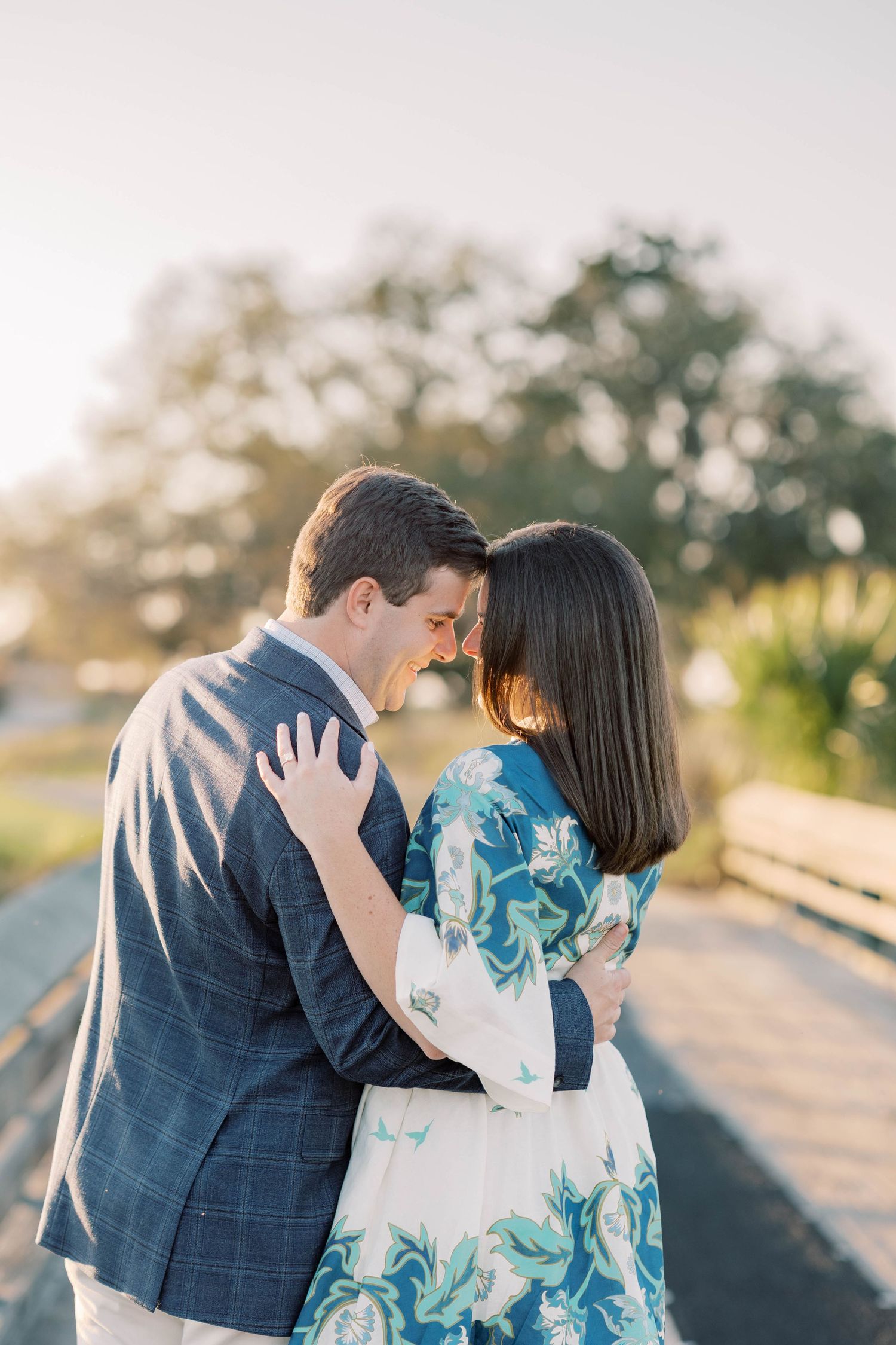 A couple shares a romantic forehead touch while embracing outdoors in golden hour light, dressed in elegant attire.