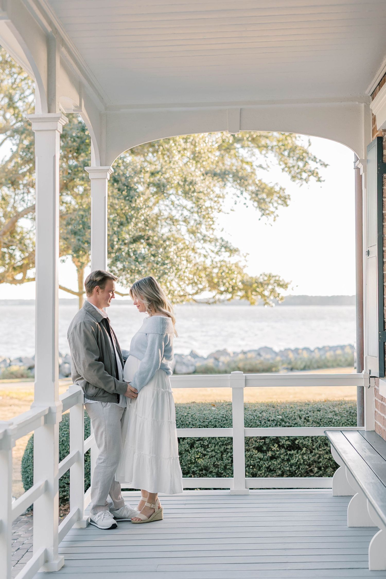 Couple sharing a tender moment on a white porch overlooking the water, woman in white dress showing a baby bump.