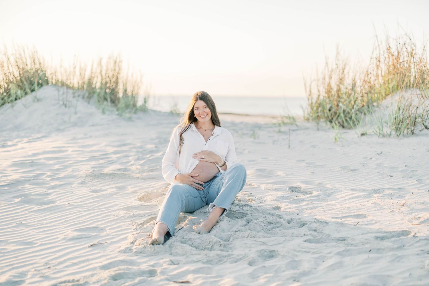 Pregnant woman sitting on sandy beach dunes in white shirt and jeans, cradling baby bump in golden sunlight.