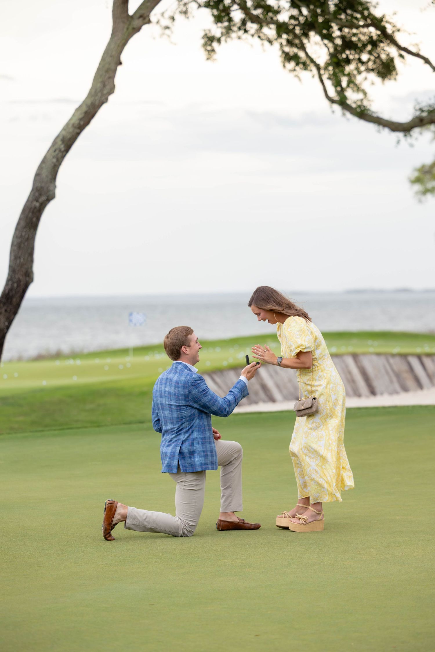 Man proposing on one knee to woman in yellow dress on golf course by the water.