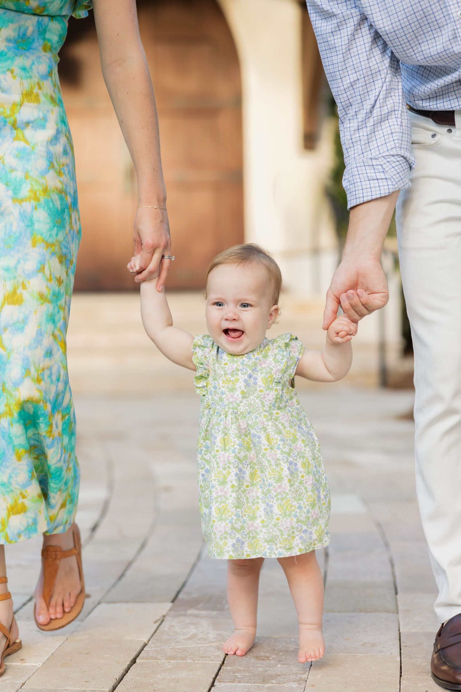 Smiling baby girl in floral dress held by parents hands learning to walk on stone pathway.