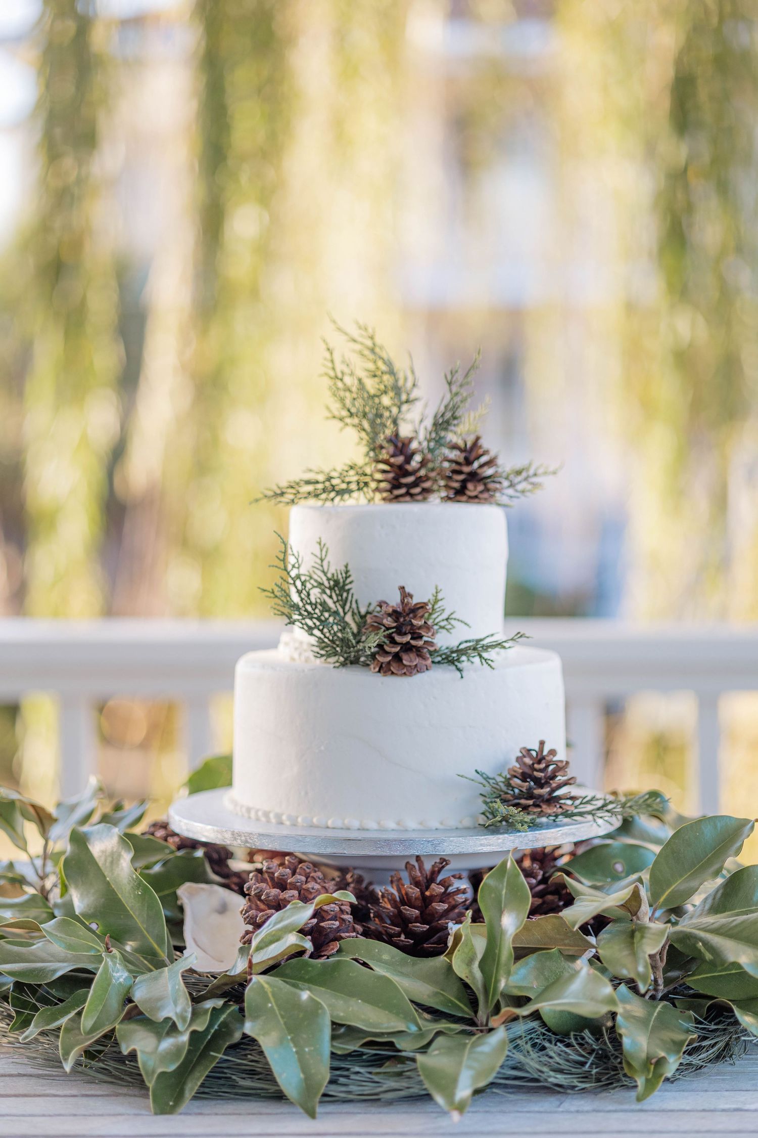 White wedding cake decorated with pine cones and greenery garland on a rustic outdoor table setting.