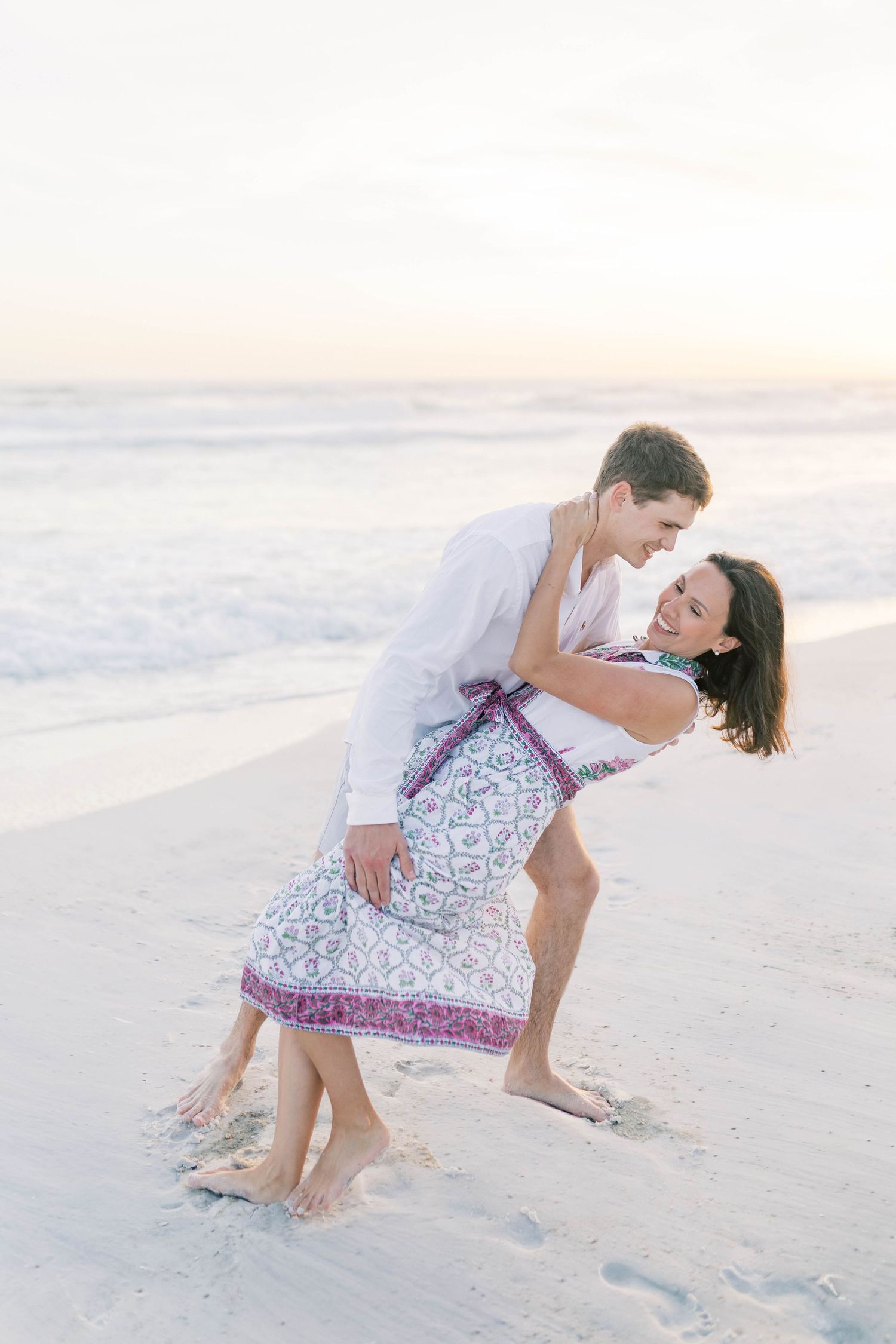 Couple sharing a romantic dip embrace on a sandy beach at sunset, woman in patterned dress, man in white shirt.