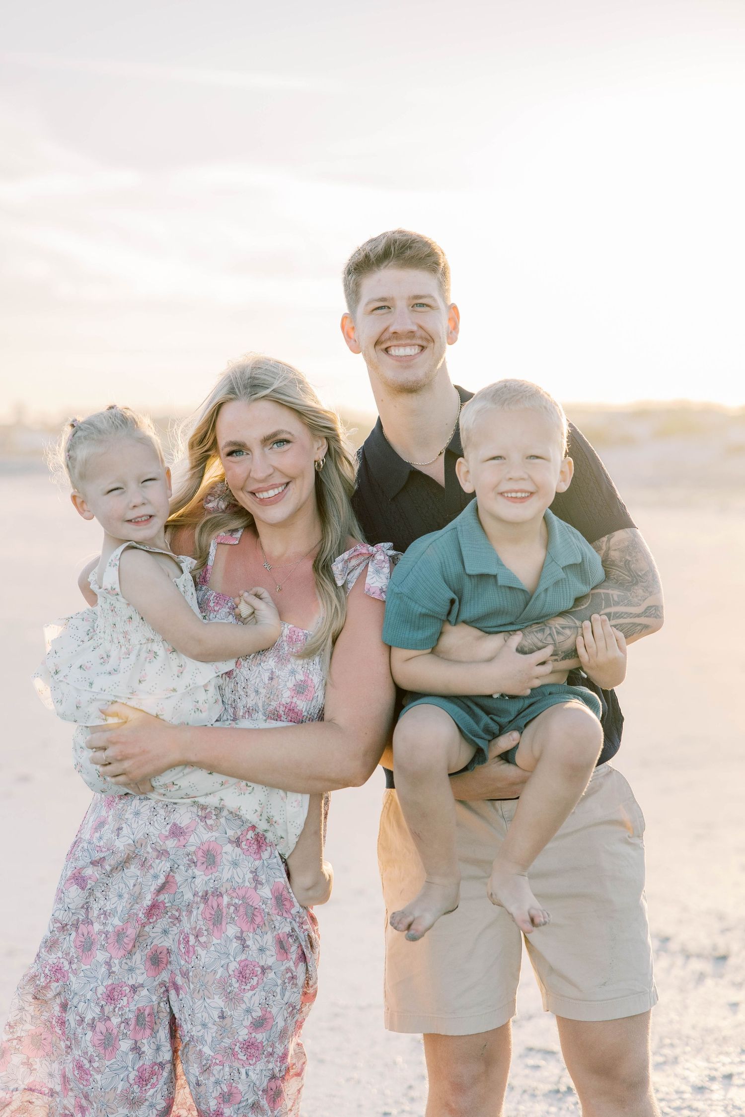 Happy family of four posing on a sandy beach at golden hour, parents holding two young children.