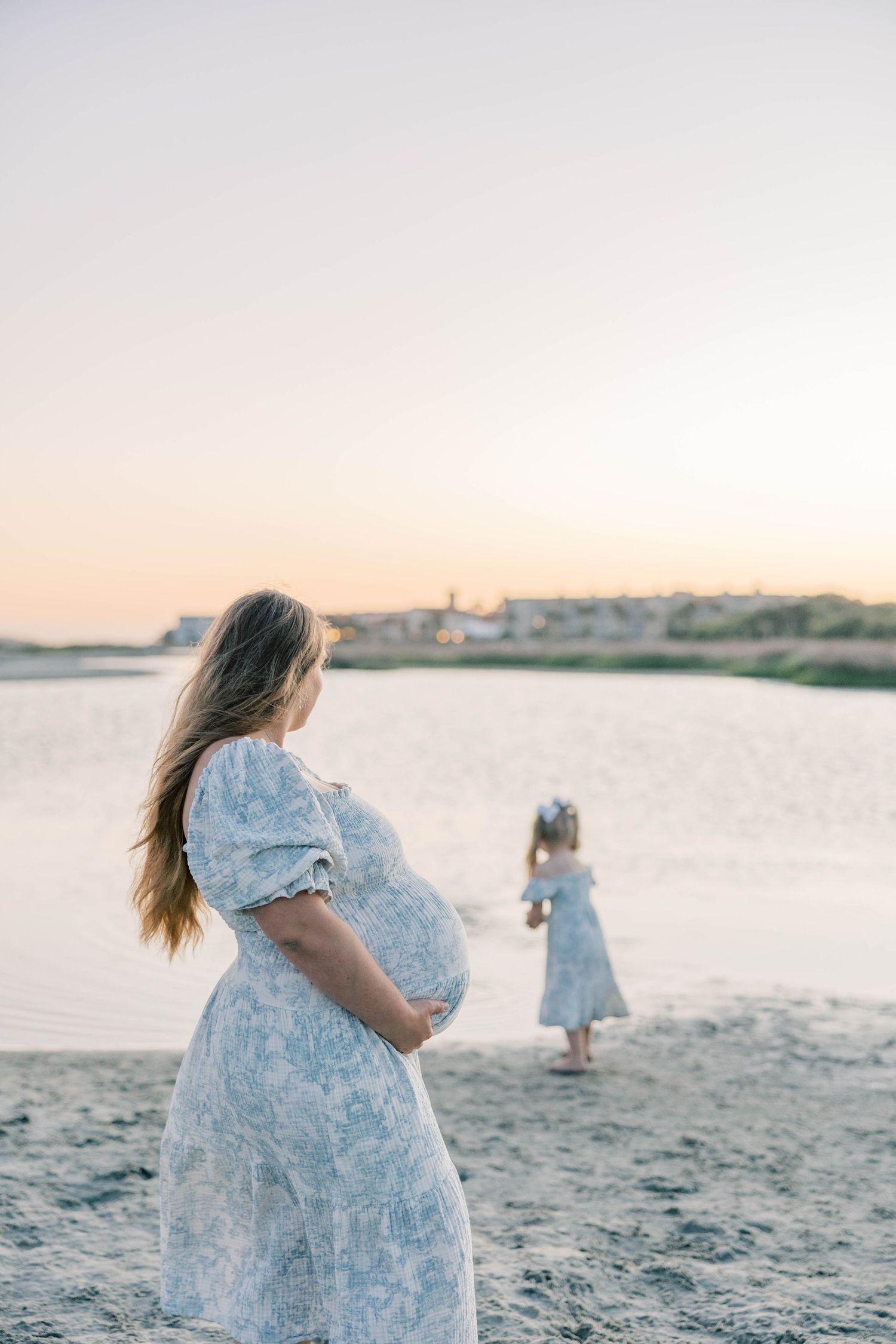 Pregnant woman in floral dress holds baby bump while young girl stands near calm water at golden hour.