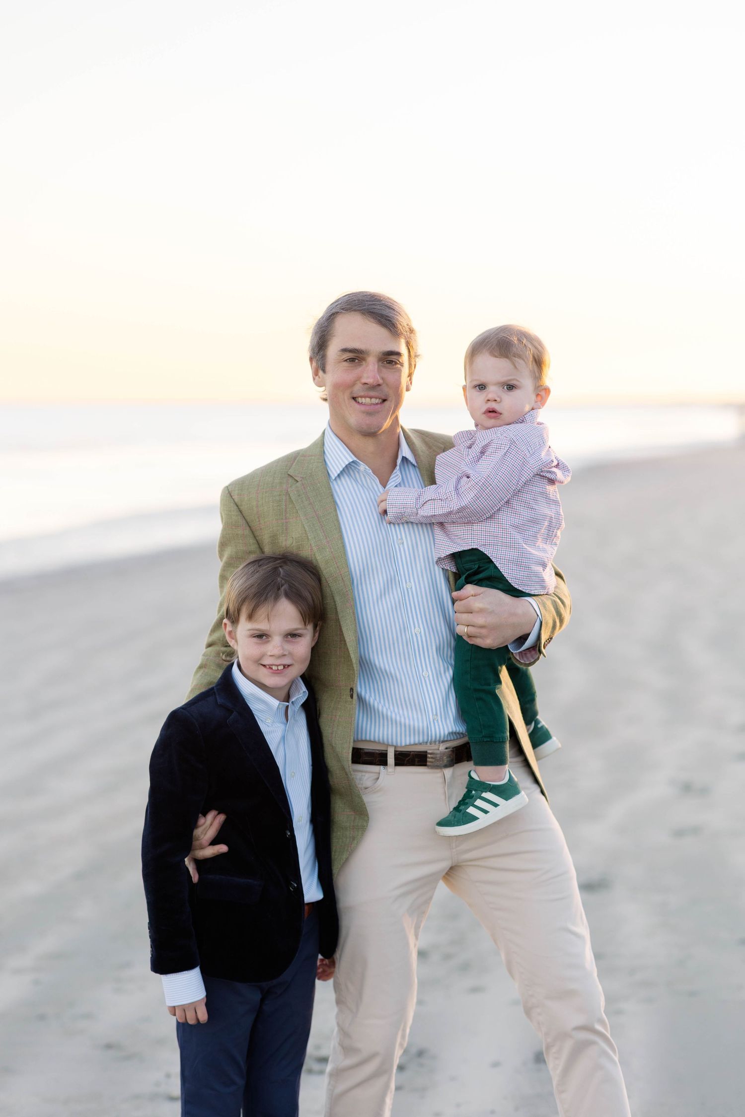 A family in coordinated beige and navy outfits poses together on a sandy beach at sunset.