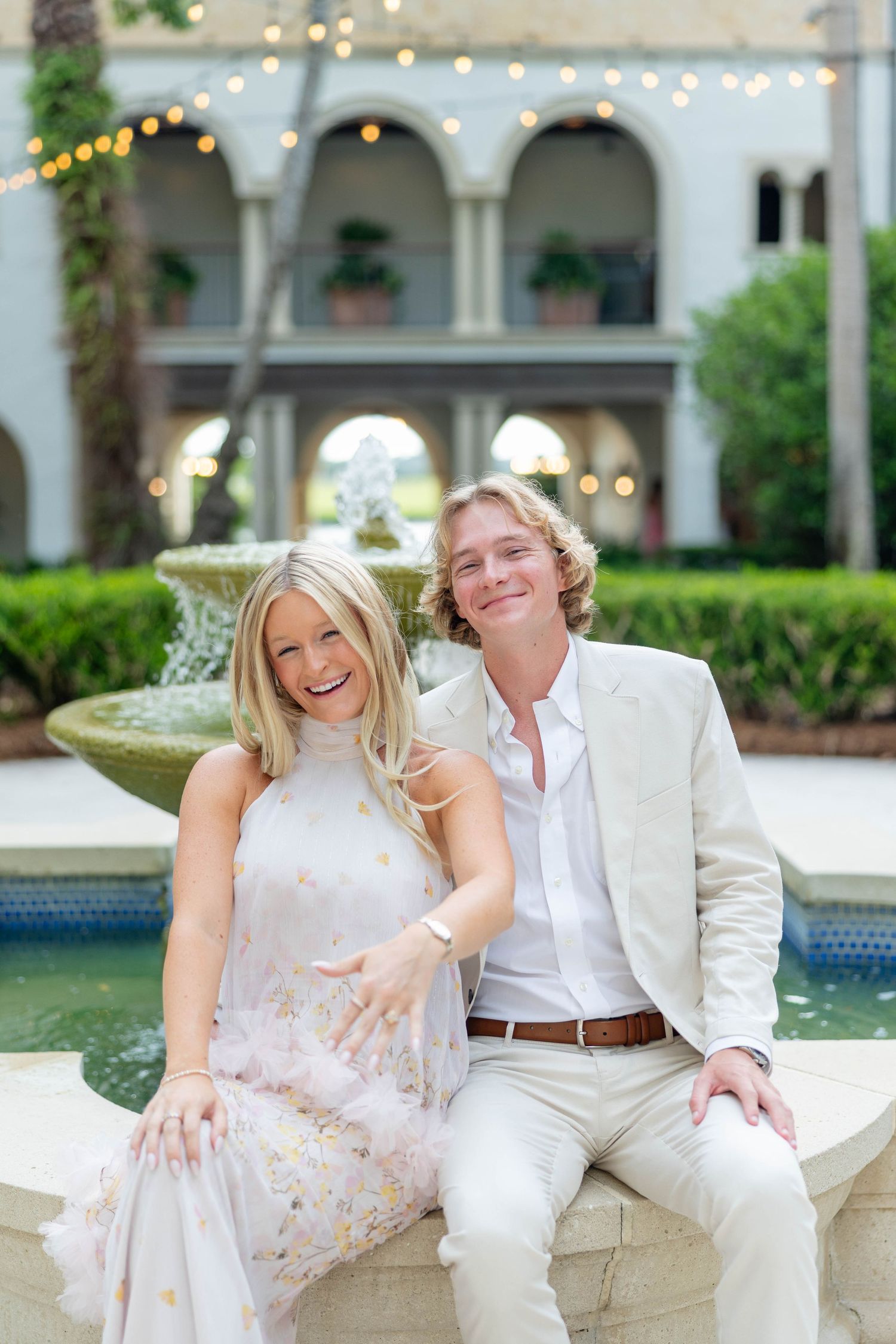 Smiling couple sitting by an elegant fountain outside a grand building adorned with string lights.