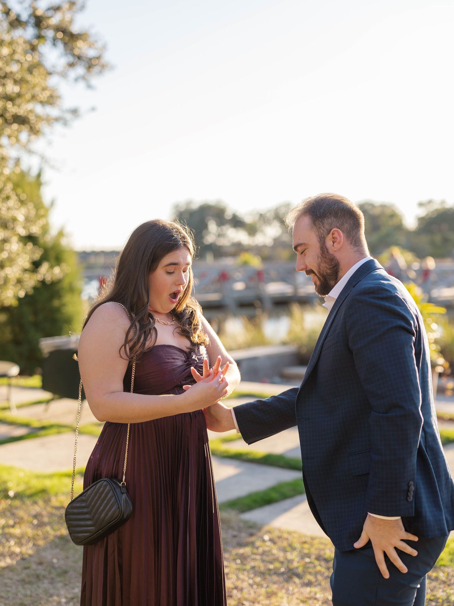 A surprised woman in a burgundy gown reacts to a marriage proposal from a bearded man in a navy suit outdoors.