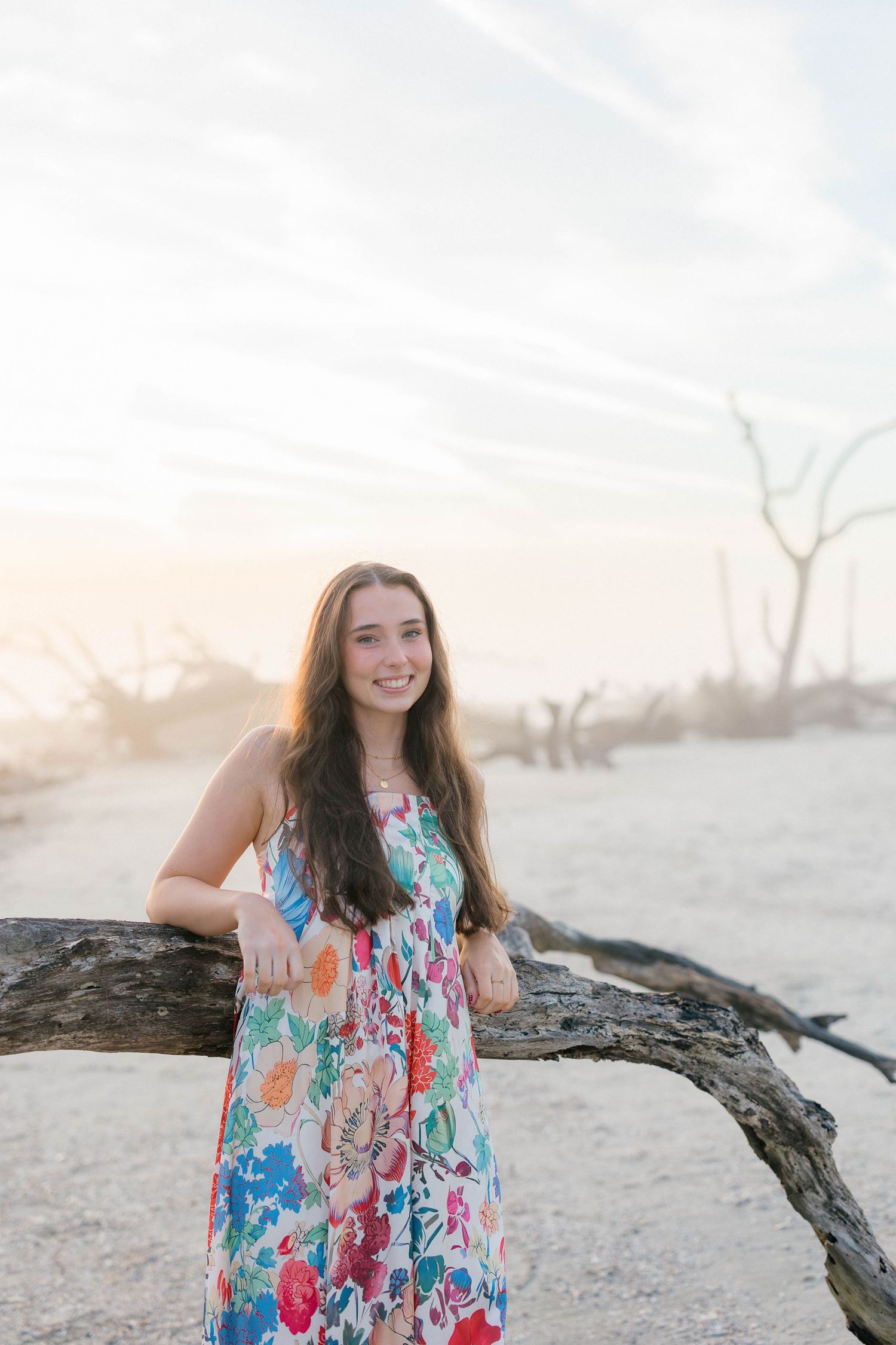 Young woman in floral dress leaning on driftwood at sandy beach during golden hour sunset.
