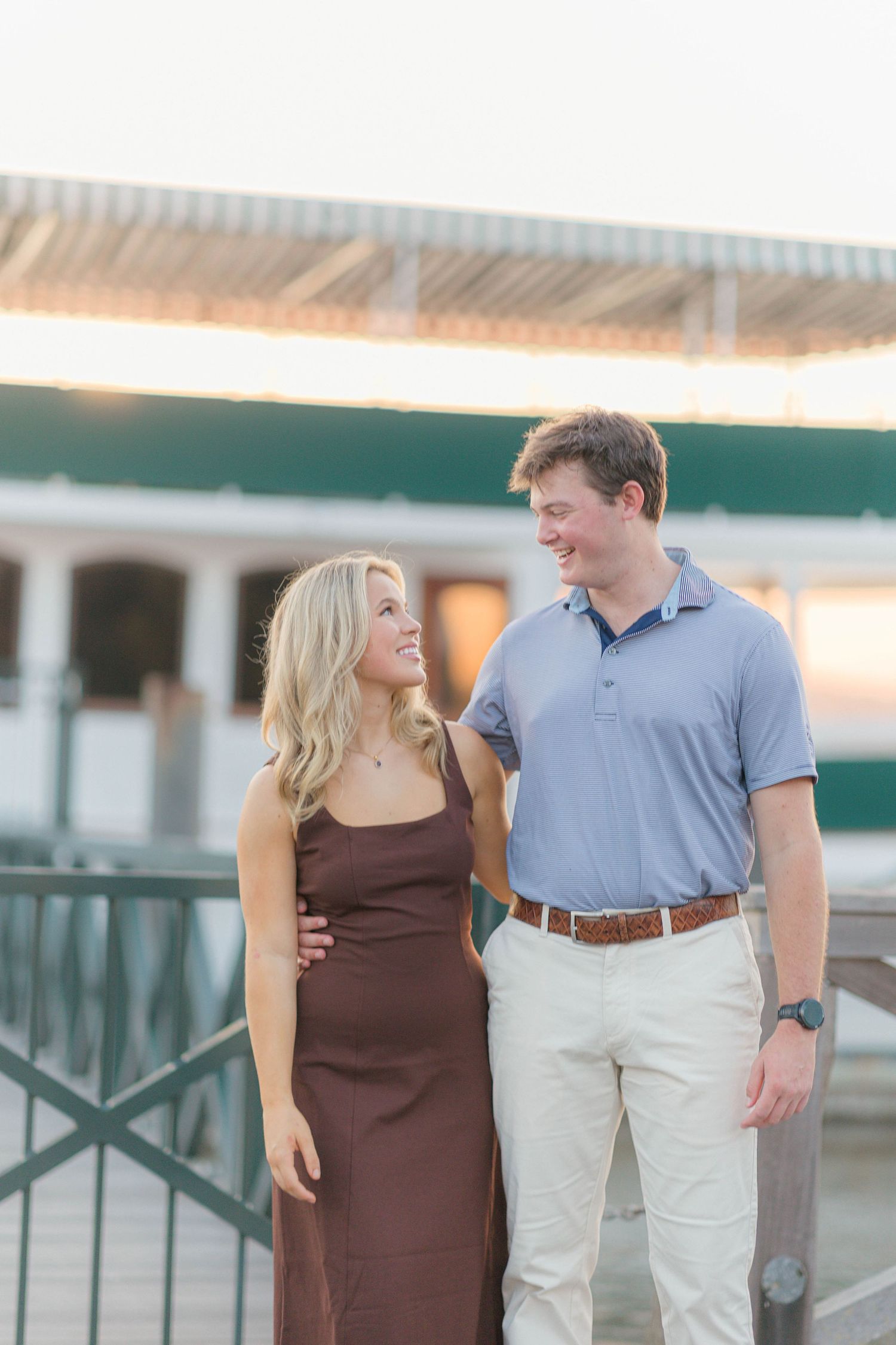 A smiling couple in casual attire share a warm moment on a waterfront dock at sunset.