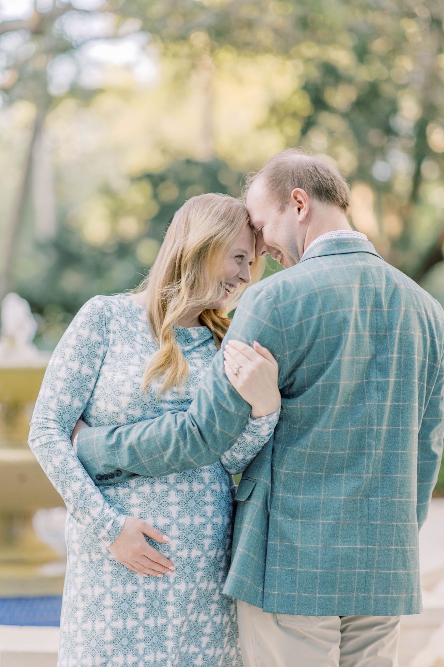 Pregnant couple in matching blue outfits sharing a tender moment outdoors, woman cradling baby bump while man leans in close.