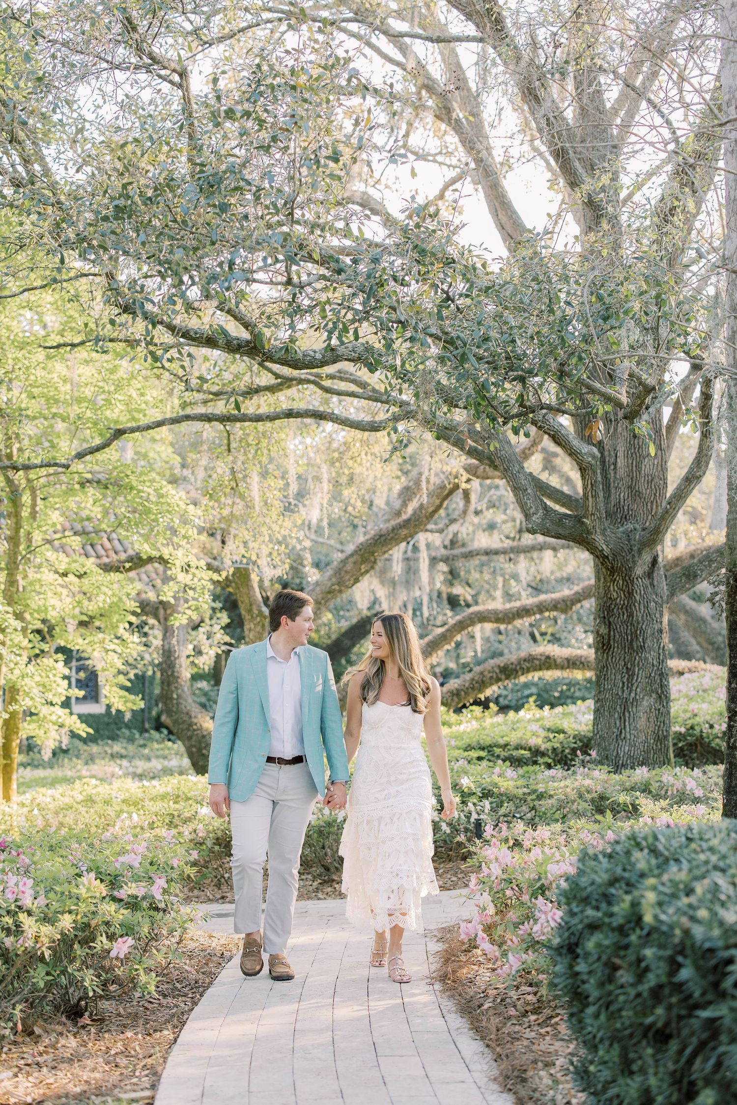 Couple holding hands walking on a brick path through a lush garden with sprawling oak trees.