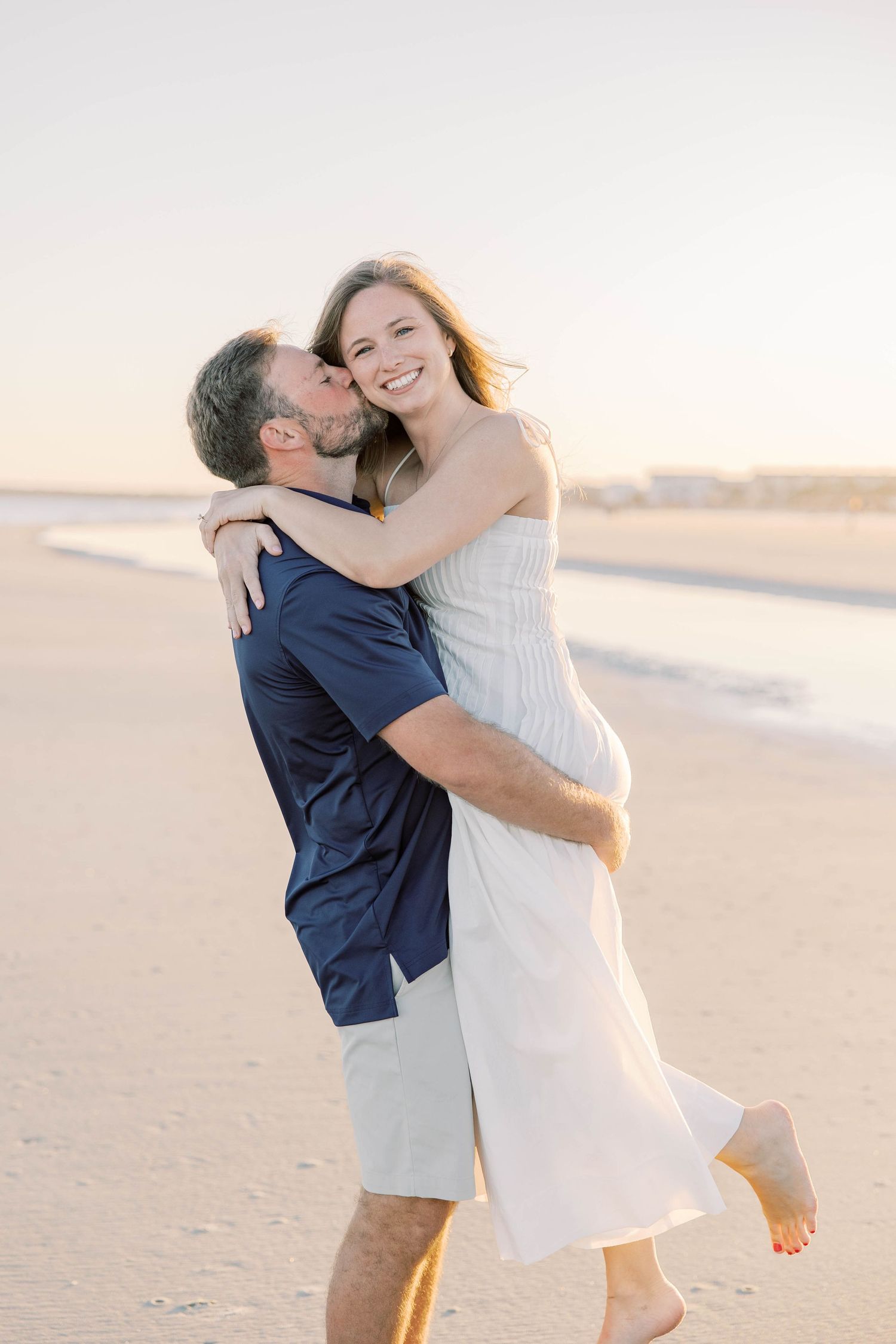 A man lifts and kisses a smiling woman in a white dress on a sunny beach at golden hour.