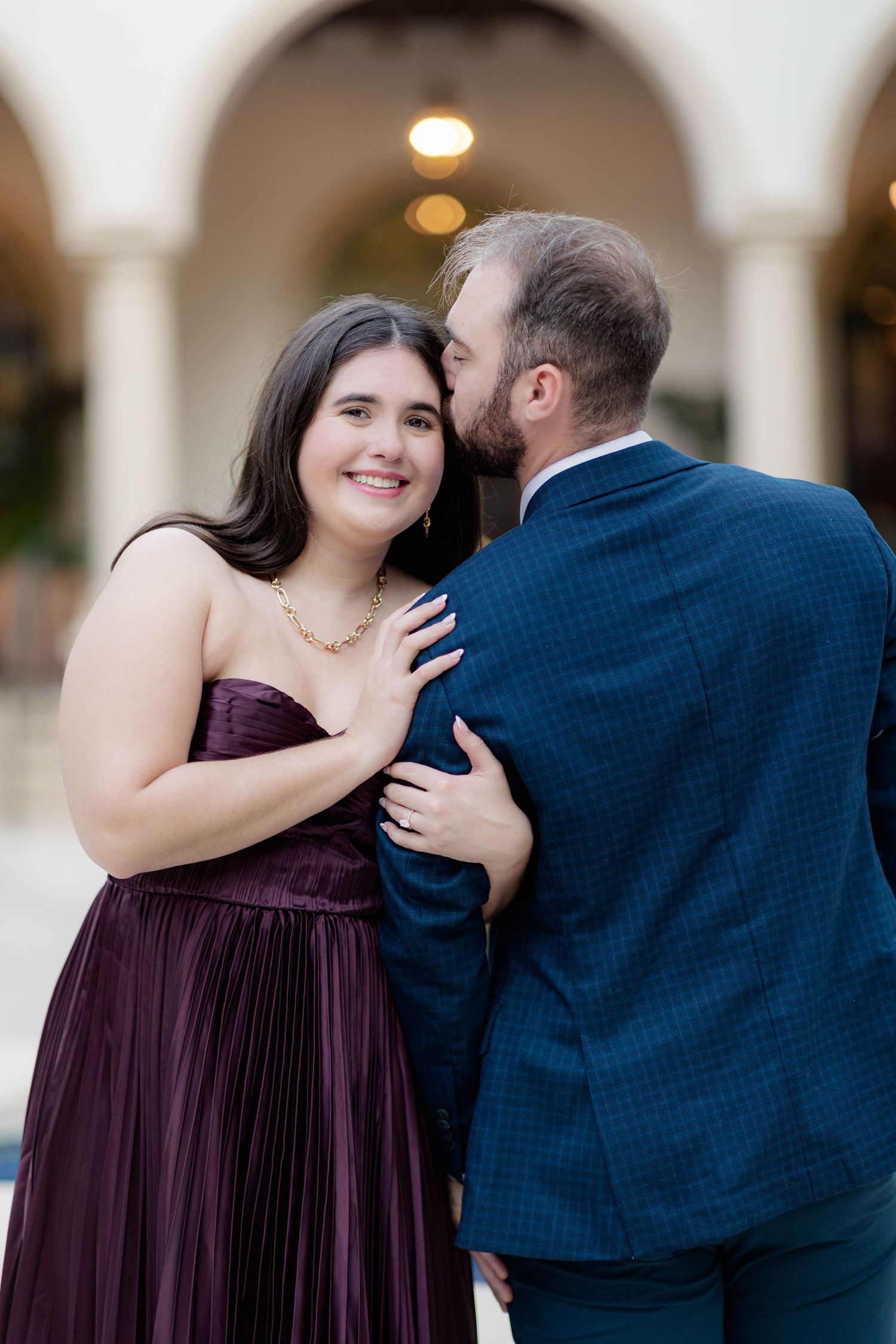 Couple in elegant attire, woman in burgundy gown smiles as man in blue suit whispers in her ear.