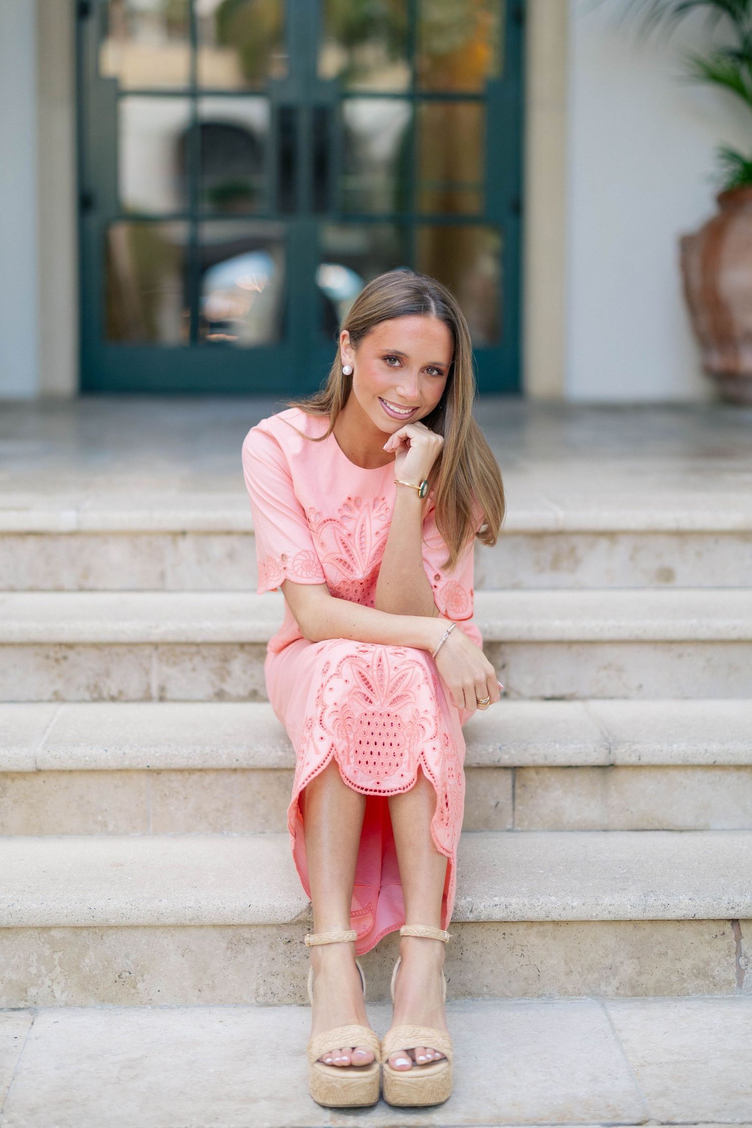 Woman in coral pink dress sitting on white stone steps with palm trees in background.