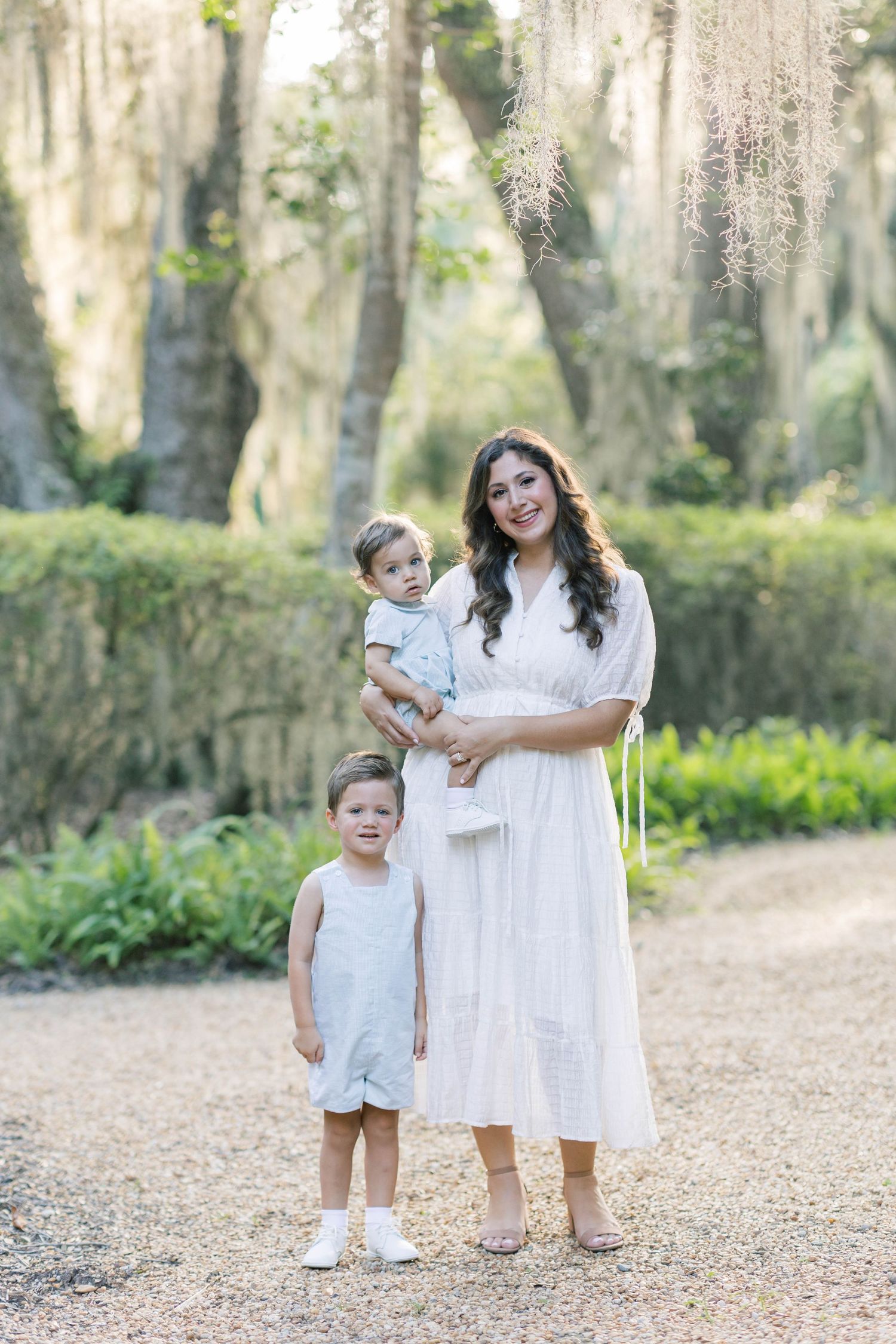 A mother in a white dress holds her baby while her toddler stands beside her on a gravel path outdoors.
