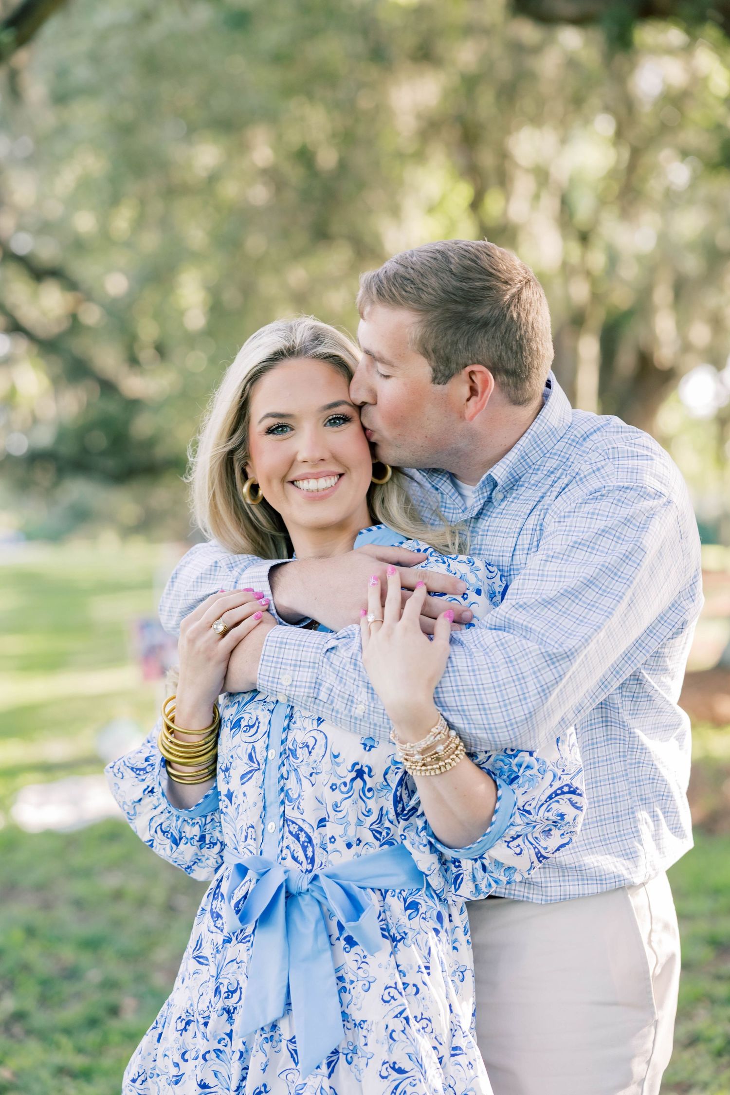 A smiling woman in a blue floral dress receives a kiss from a man in a plaid shirt outdoors.