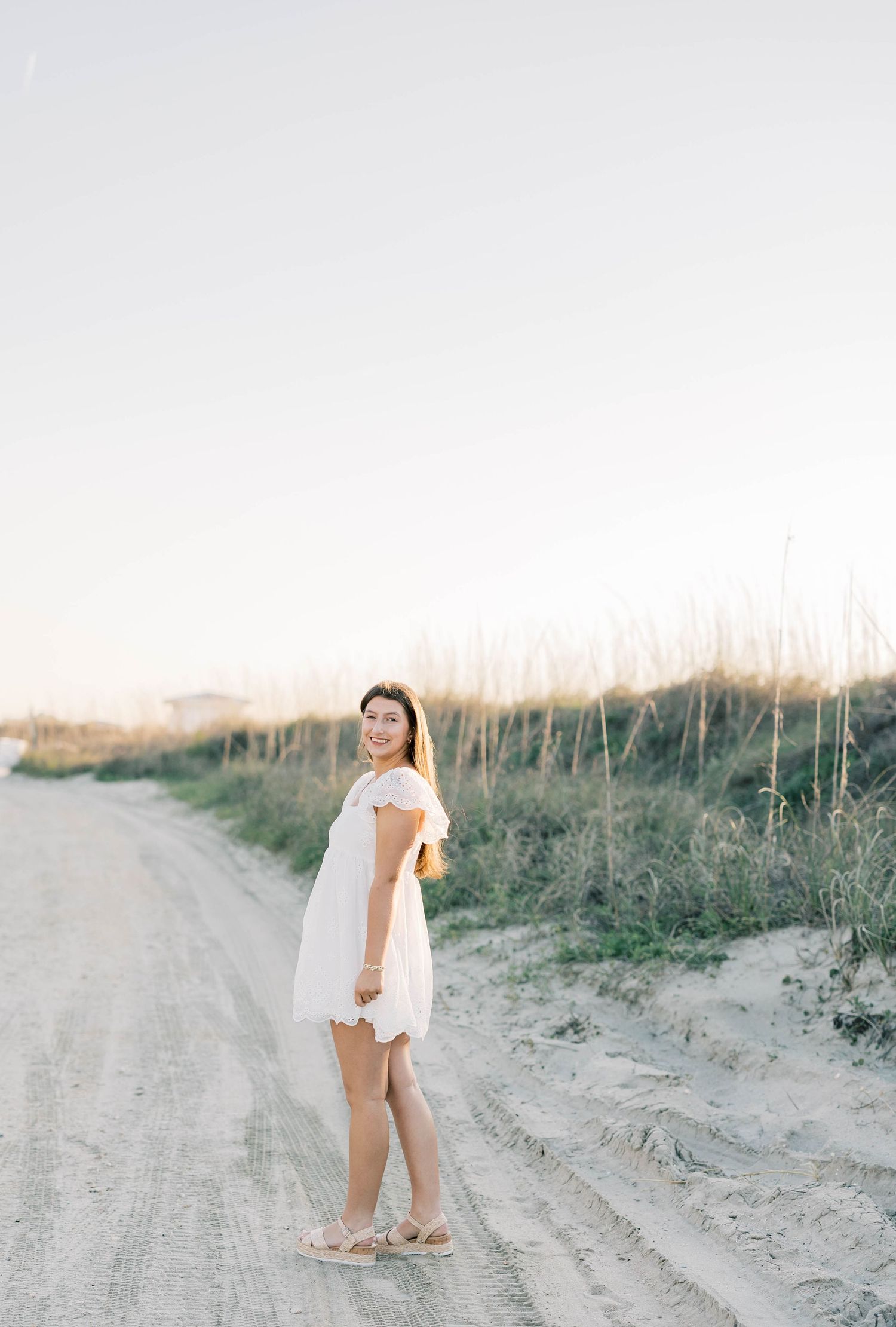 Young woman in white mini dress smiling on sandy beach path at golden hour.